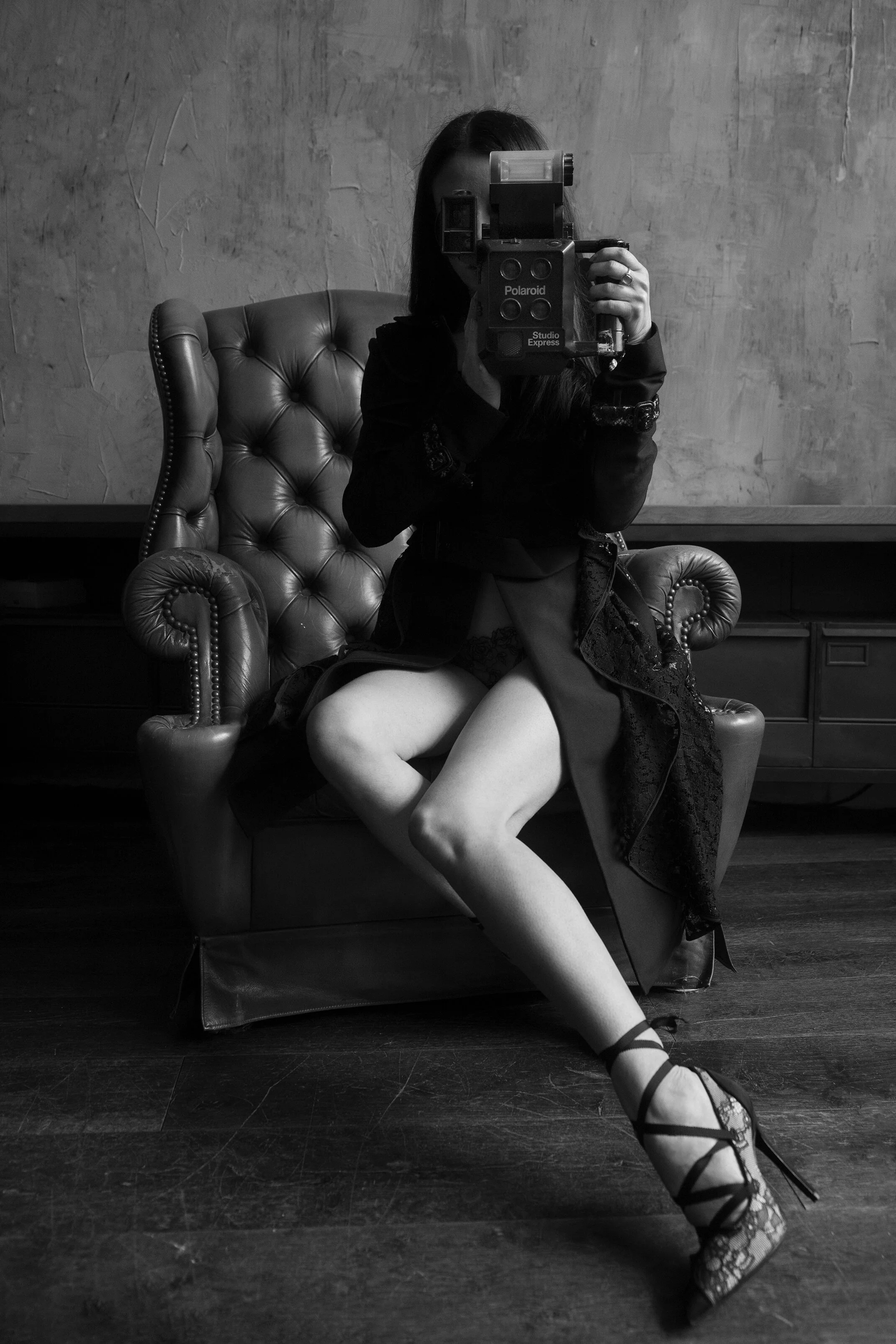 Une femme assise sur un fauteuil en cuir, portant des talons à lacets, prenant une photo dans un miroir, en noir et blanc.