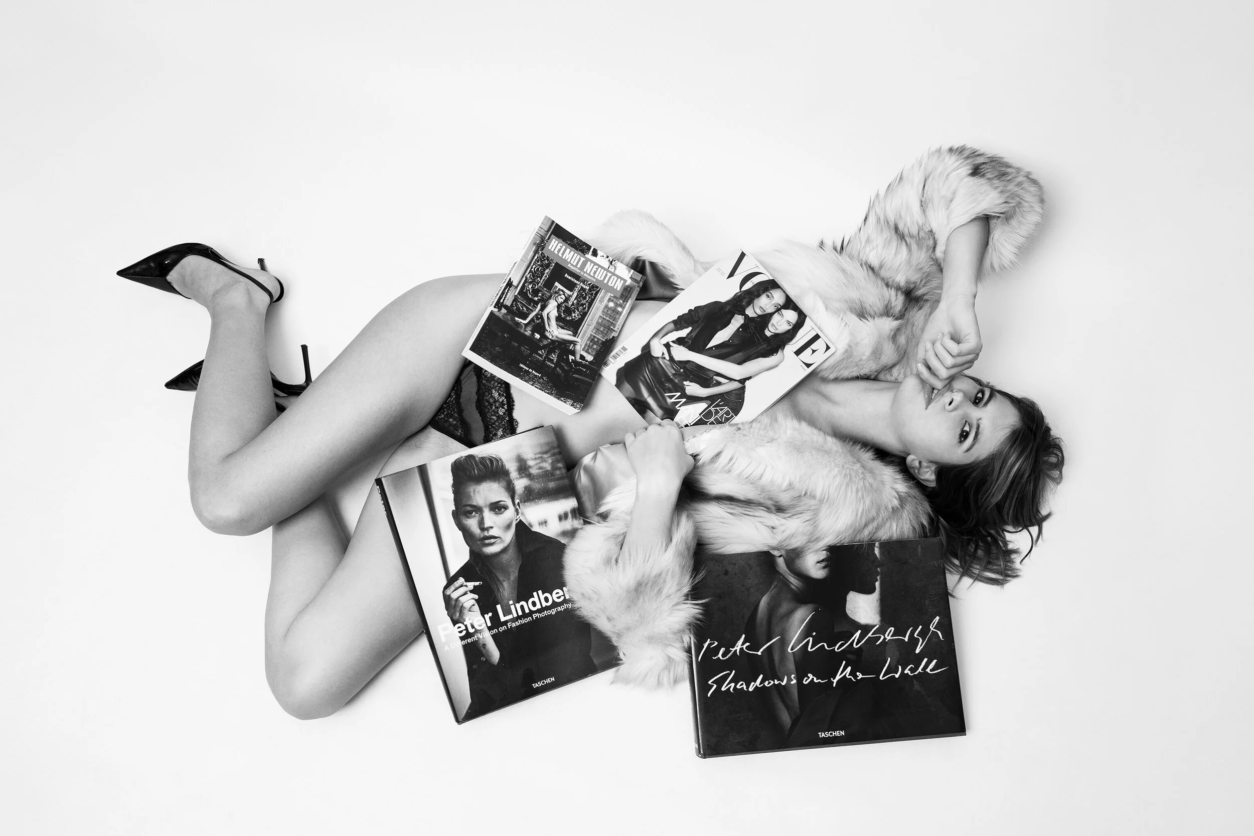 Femme allongée sur le sol, entourée de magazines de mode, portant un manteau en fourrure et des chaussures à talons, en noir et blanc.