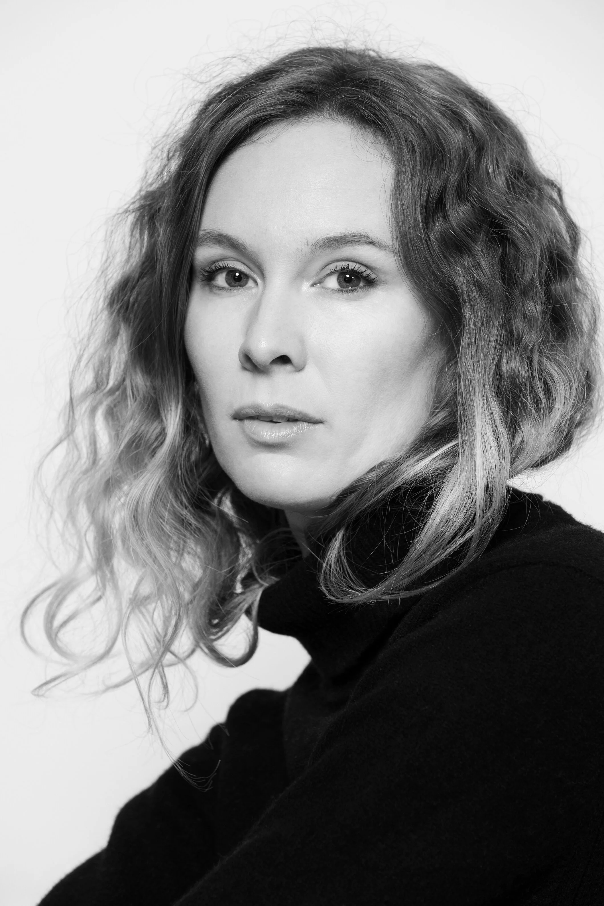 Portrait noir et blanc d'une femme avec des cheveux bouclés, portant un pull noir, regardant vers l'appareil avec une expression neutre.