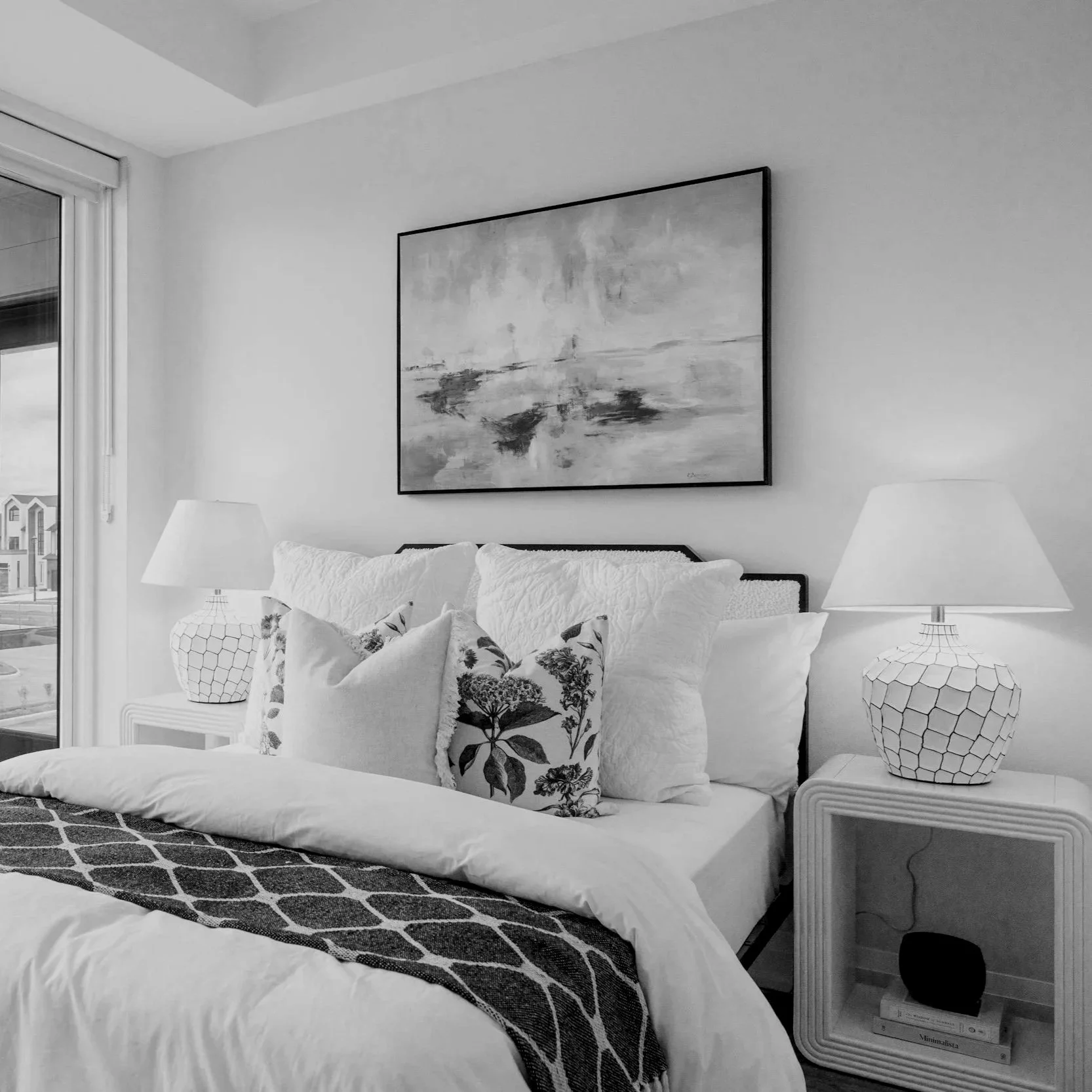 A bedroom with a bed featuring multiple pillows, two bedside tables with lamps, a painting on the wall, and a window with blinds showing neighboring houses outside.