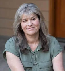 A woman with long gray hair wearing a green shirt, smiling softly inside a room with wooden furniture.