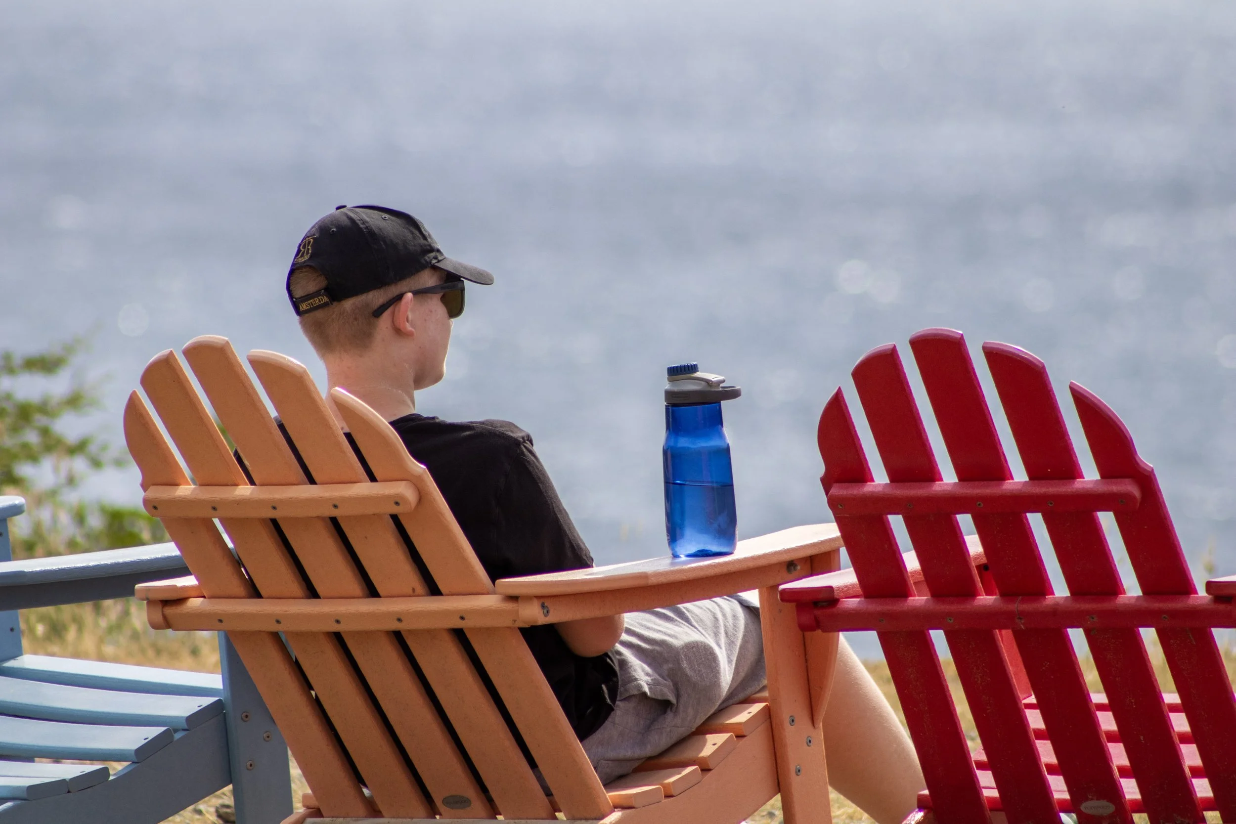 A young person sitting on a wooden chair by the water, wearing sunglasses and a black cap, with a blue water bottle on a small table in front of them, facing away from the camera.