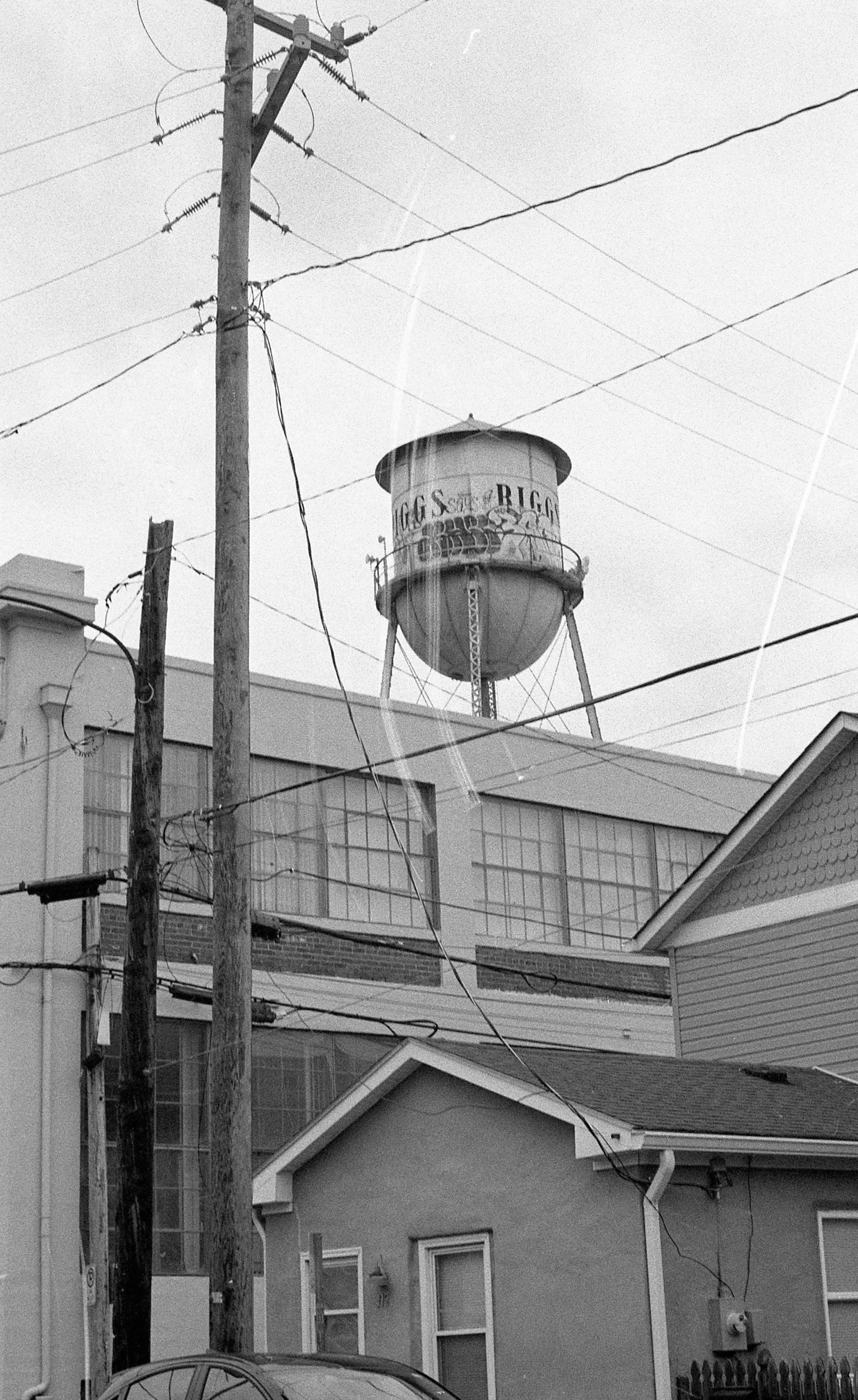 A black and white photo of a water tower with graffiti, utility poles, and power lines in a neighborhood.