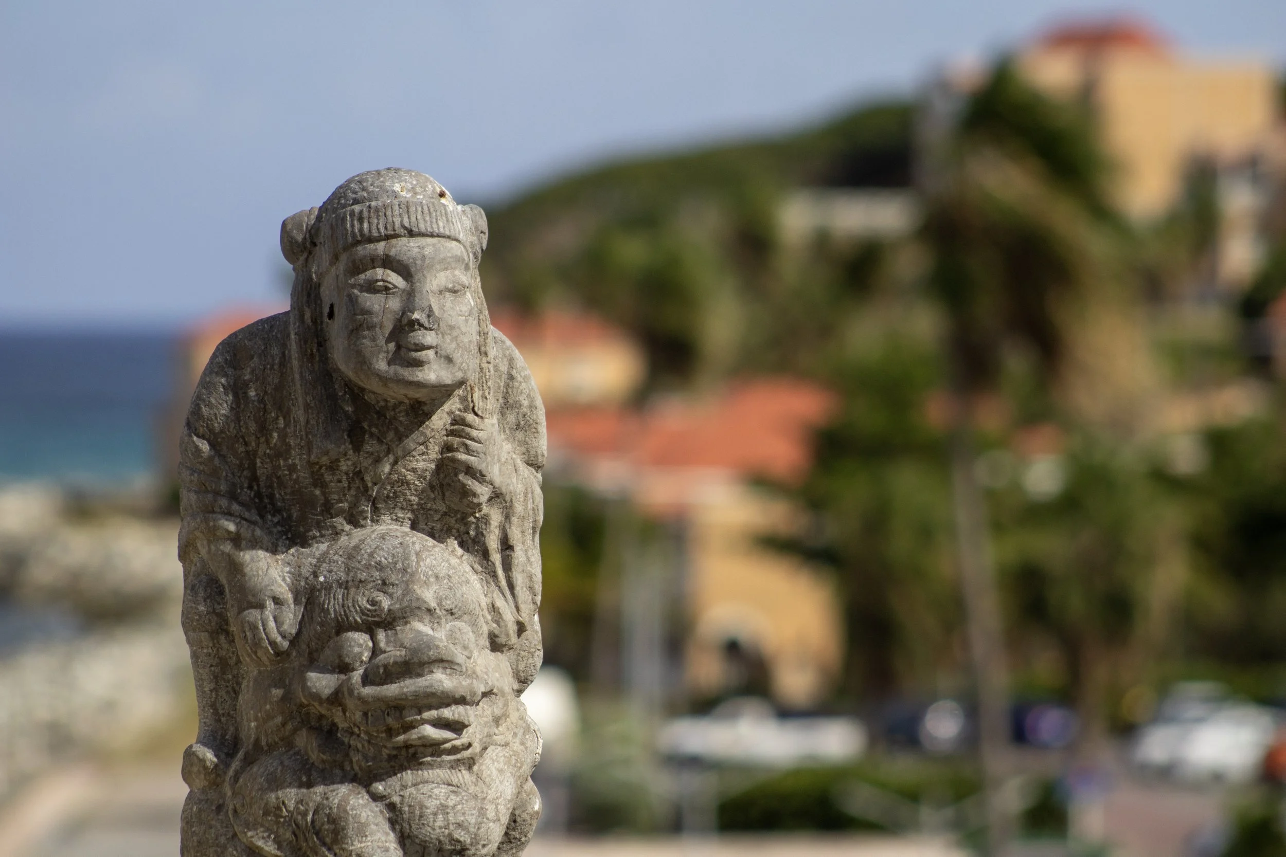 Close-up of ancient stone sculpture depicting a figure with a headdress, holding an object, with a blurred background of buildings, trees, and a water body.