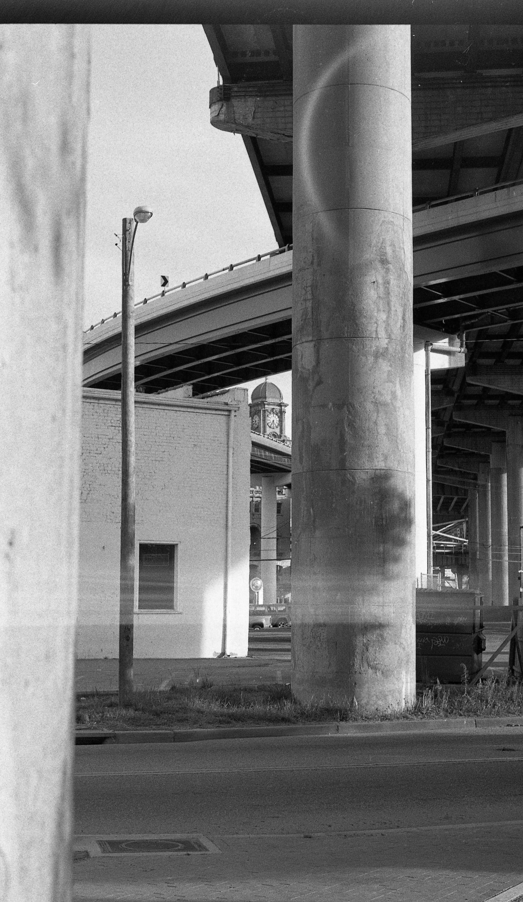 Black and white photo of large concrete pillars supporting an elevated highway, with a streetlamp, brick building, and clock tower in the background.