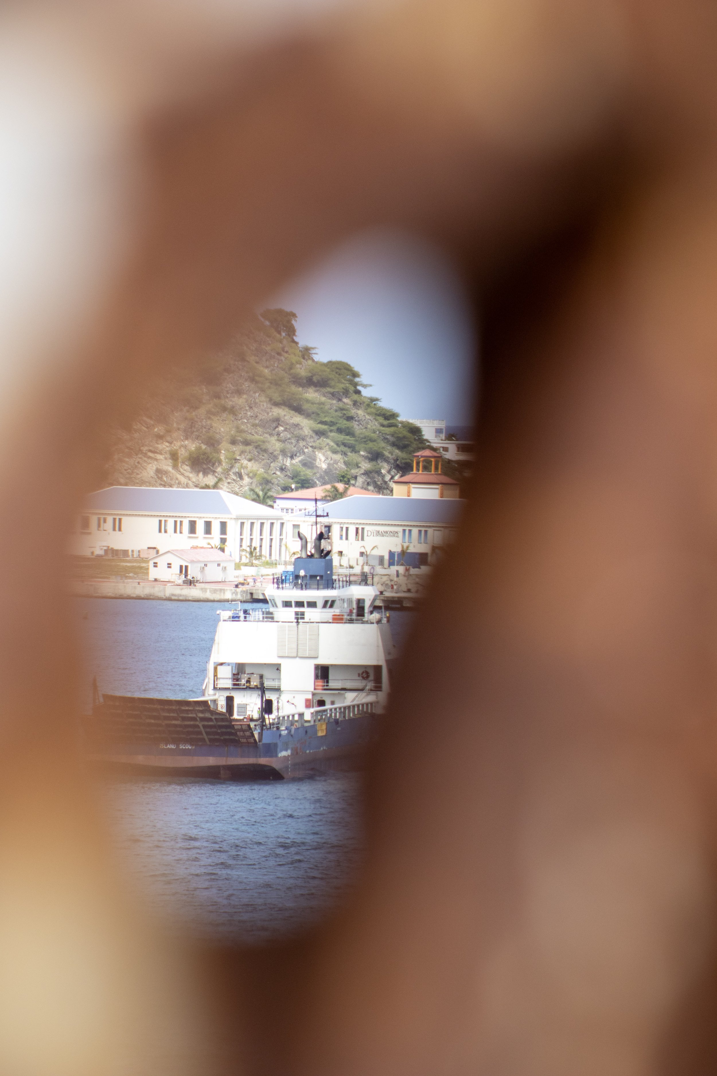 A ship seen through a circular opening in a wooden structure, with buildings and a hillside in the background.