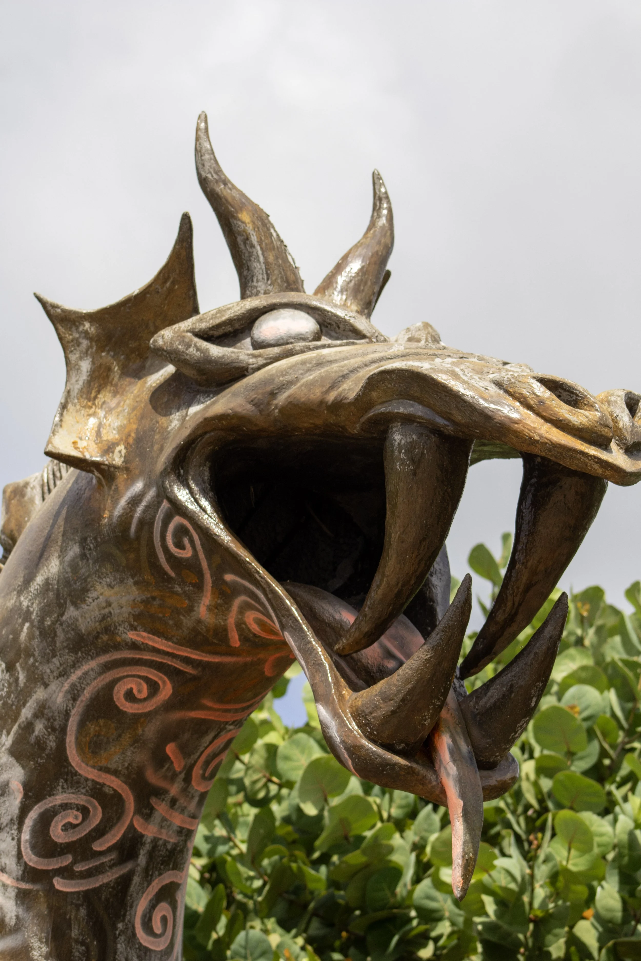 Close-up of a dragon sculpture with an open mouth, made of metal with intricate patterns, set against a background of green bushes and a cloudy sky.