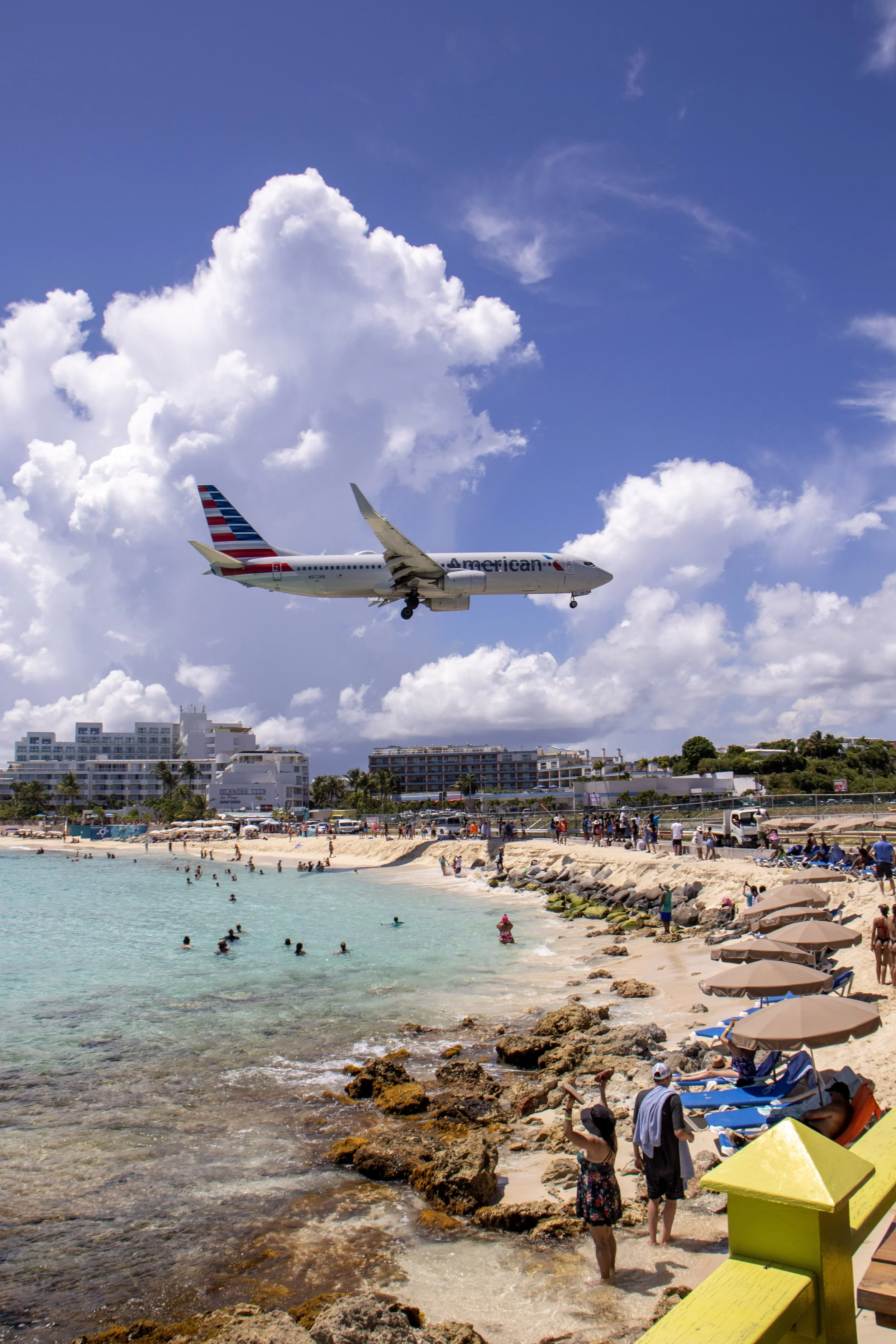 An airplane flying low over a beach with people swimming in the water and sunbathing on the sand, with buildings and blue sky with clouds in the background.