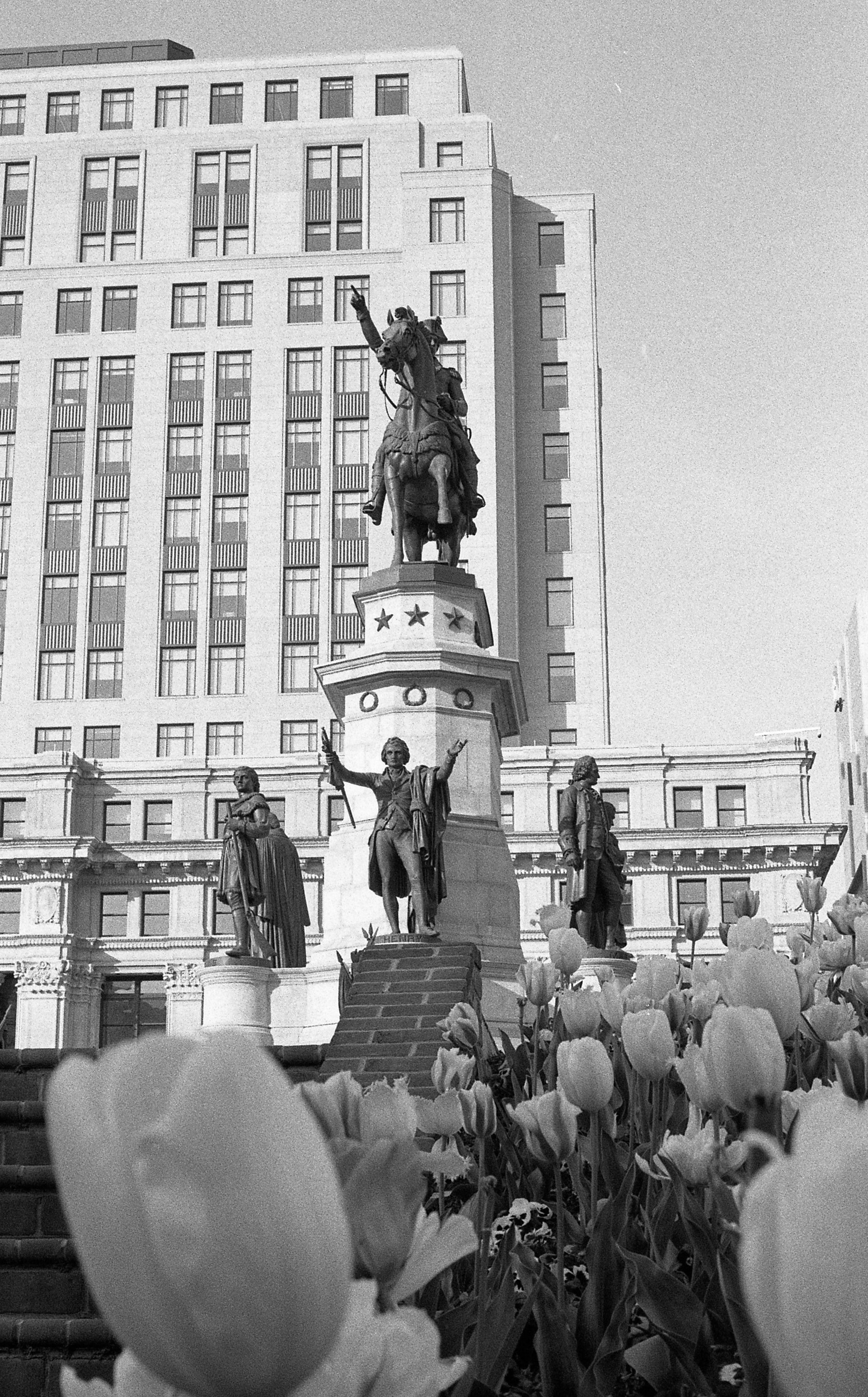 Black and white photo of a monument with a statue of a man on a horse at the top, surrounded by four statues of people on pedestals, with a tall building in the background, and flowers in the foreground.