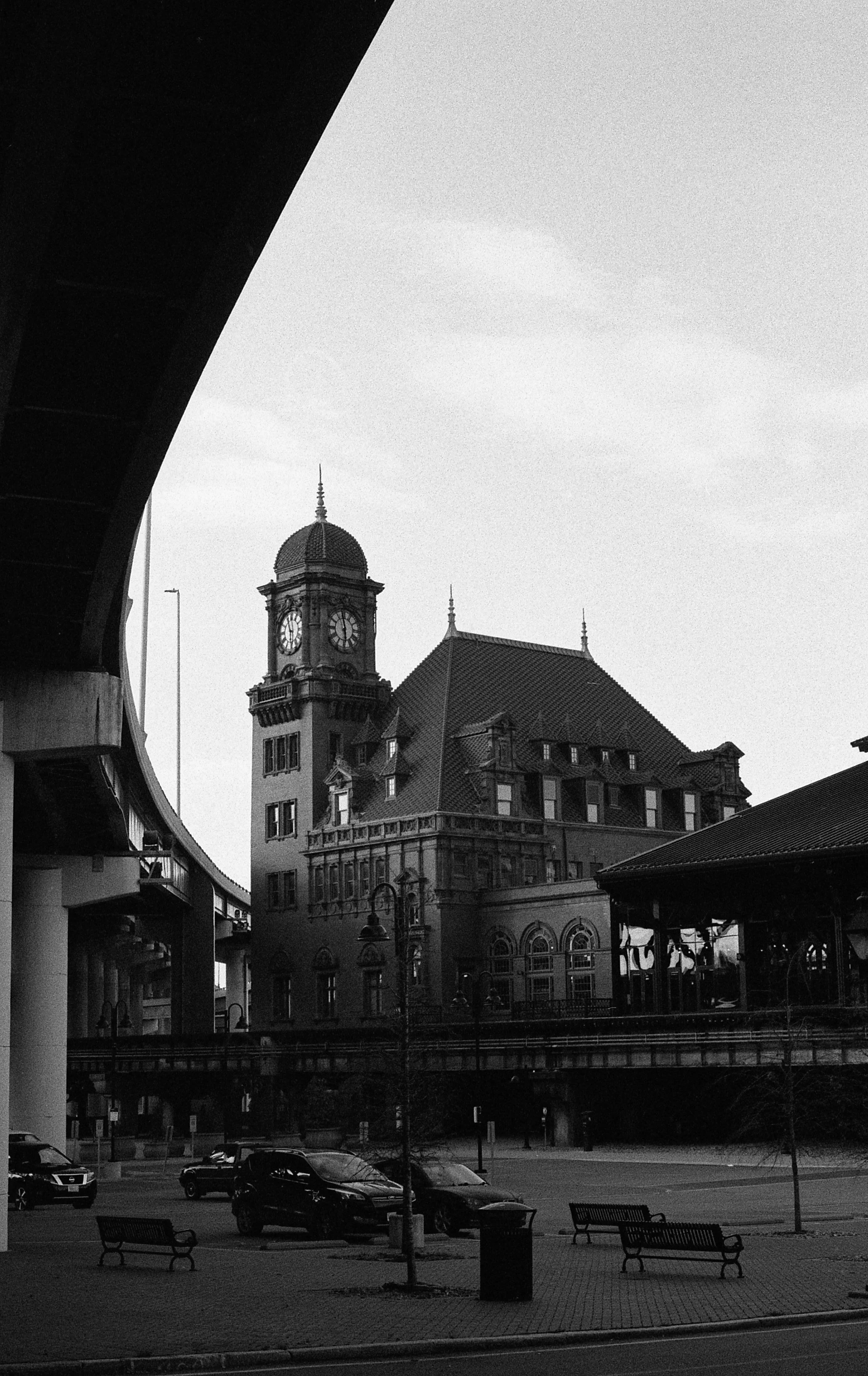 Black and white photo of a historic clock tower and a large building with a steep roof, shot from beneath an arched overpass. Cars are parked nearby, with benches and lampposts in the foreground.