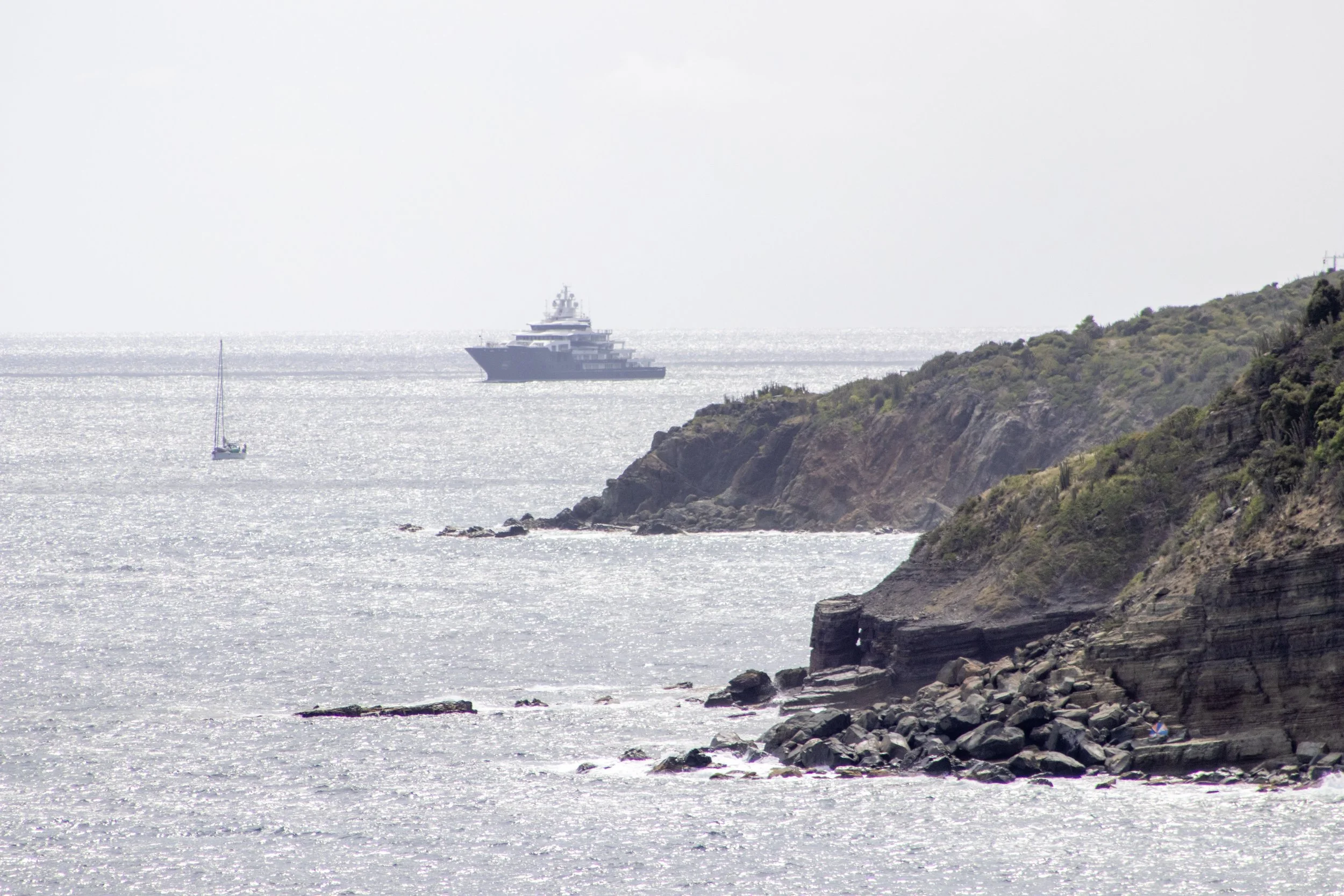 View of the ocean coastline with cliffs on the right and boats in the water, including a large yacht and a smaller sailboat.
