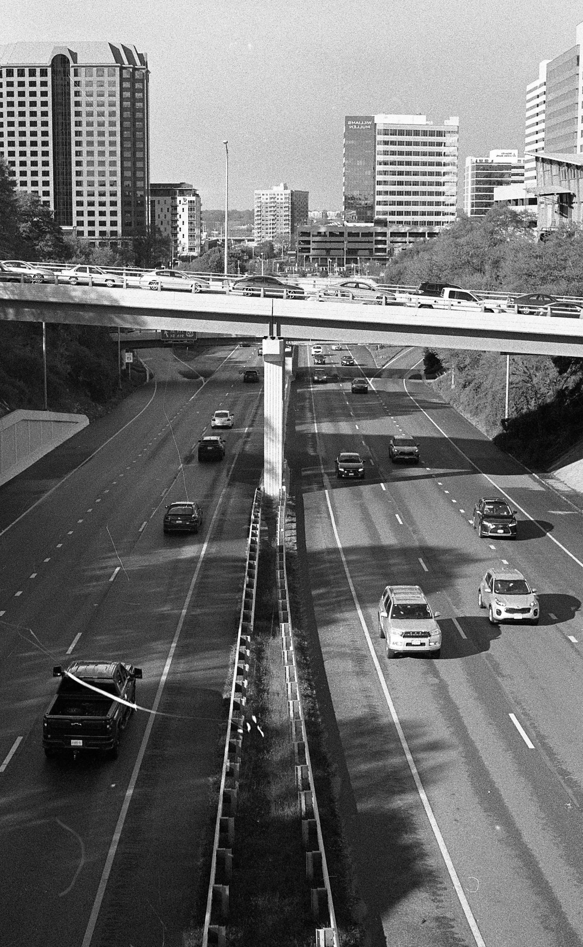 Black and white image of a city highway with cars traveling in both directions, overpass with additional cars, and tall city buildings in the background.