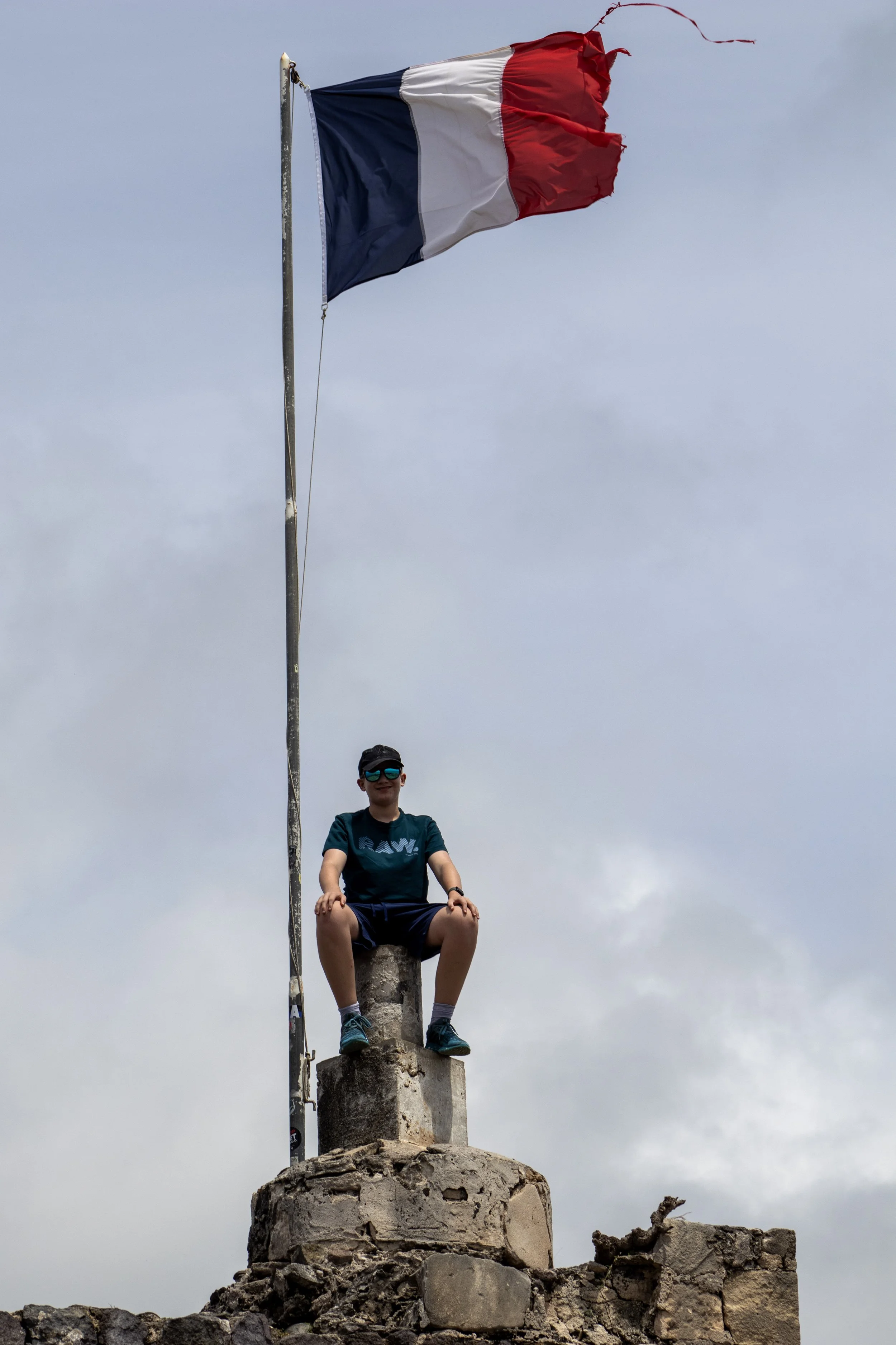 A person sitting on a stone monument with a large French flag flying high behind him, on a cloudy day.