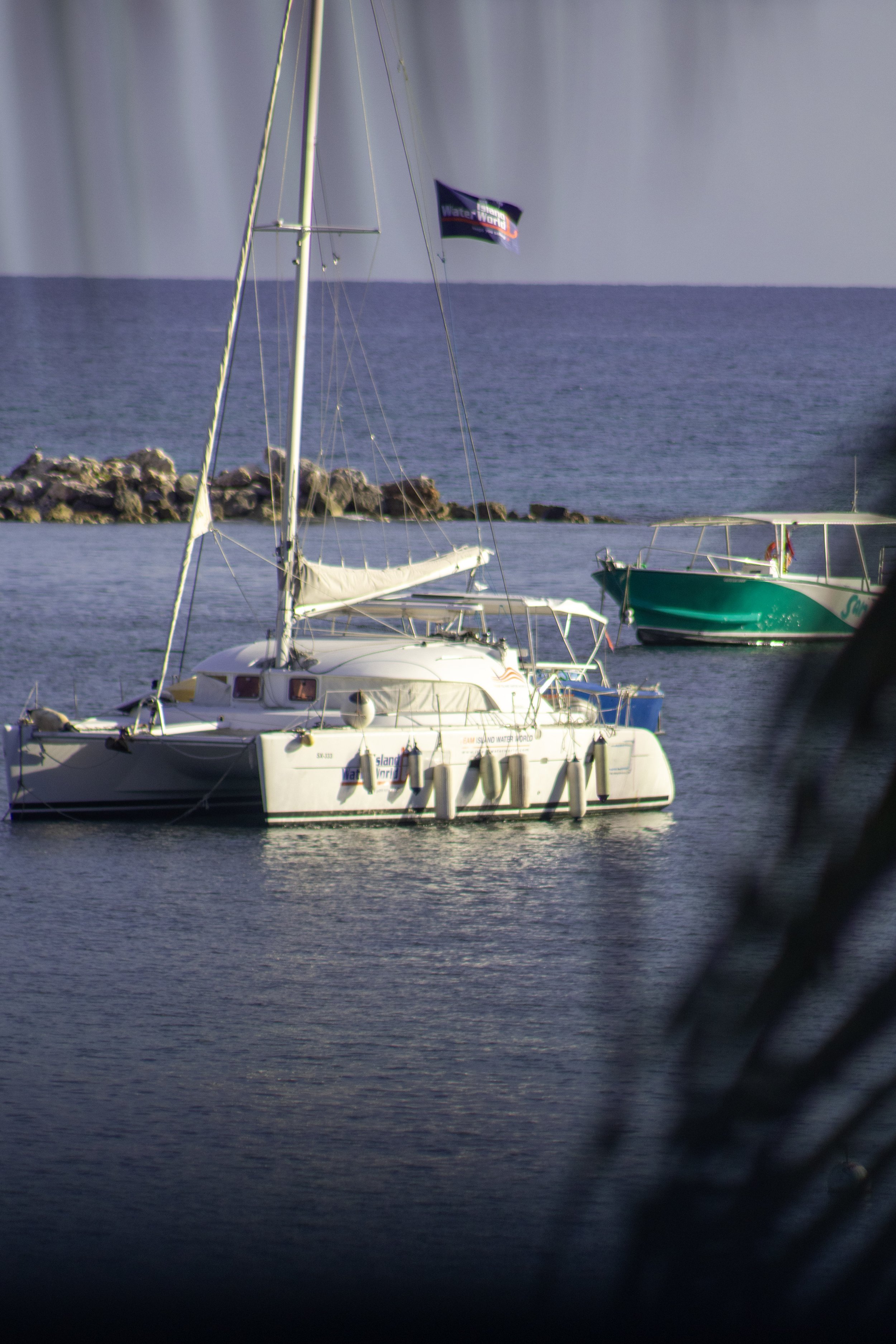 View of sailboats and motorboats anchored in a calm body of water, with a rocky breakwater in the background and a flag reading 'Water World' on one boat.