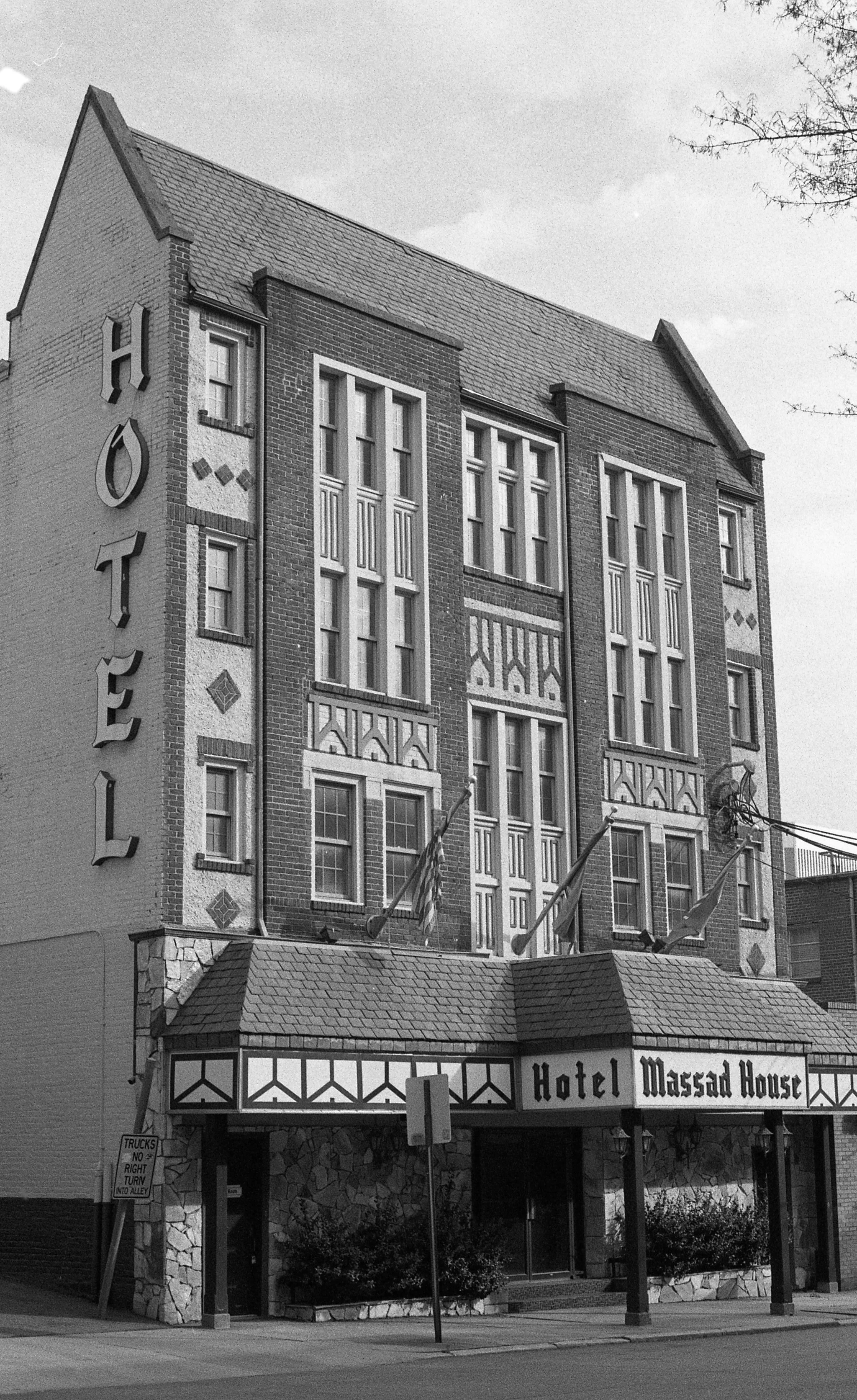 Black and white photo of the Hotel Massad House building, featuring a multi-story brick facade with decorative window designs, pointed rooflines, and flags hanging from the entrance canopy with a stone exterior base.