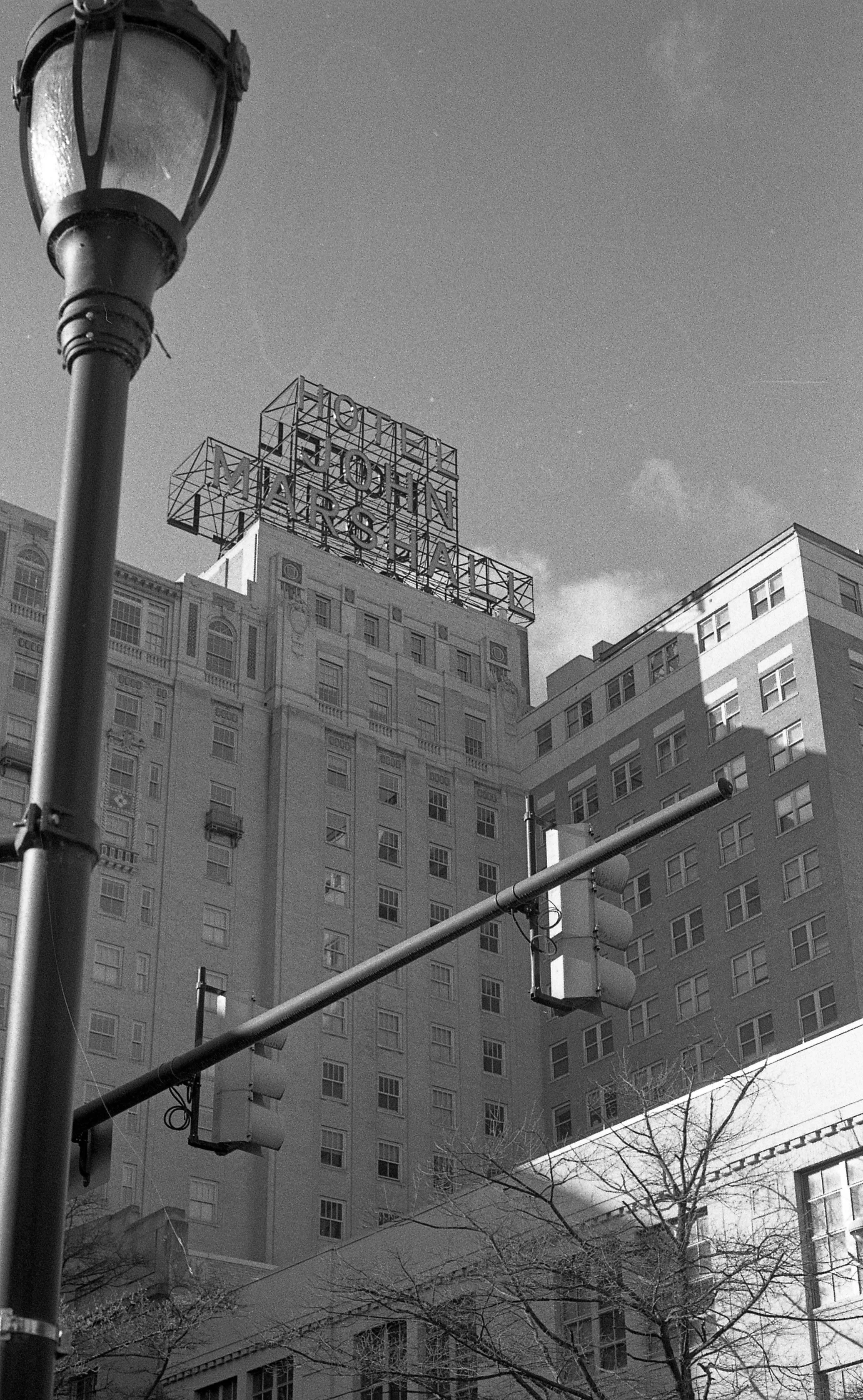 Black and white photo of the Hotel John Marshall building with an illuminated sign on top, street lamp in the foreground, traffic lights, and leafless trees at the bottom.