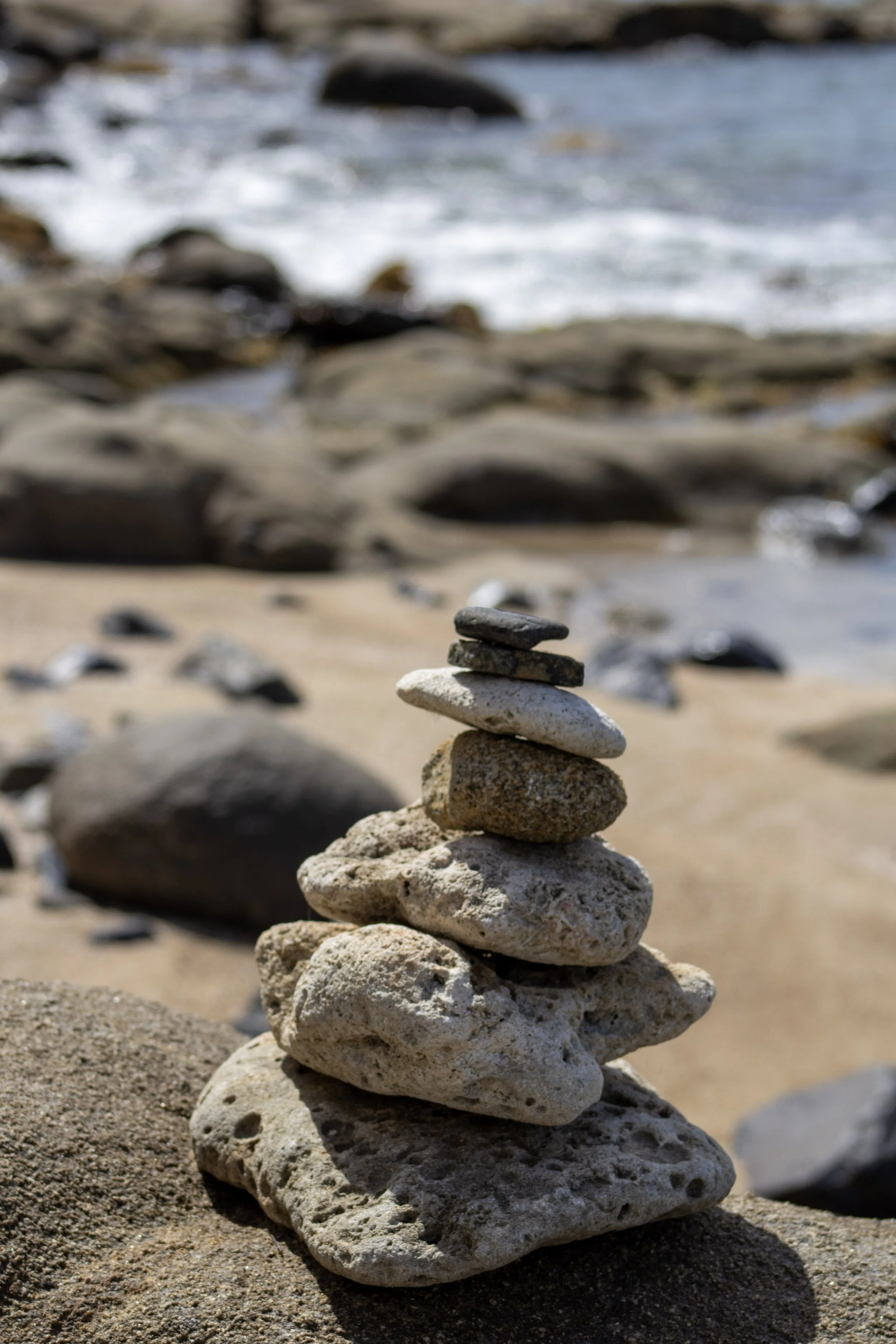 Stack of seven smooth stones of varying sizes and colors balanced on a sandy beach with rocks and ocean waves in the background.