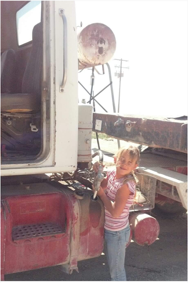 A young girl standing next to a large, old, red truck, holding onto the truck's door handle and smiling at the camera. The truck appears weathered, with a large, rusty tank and exposed mechanical parts. Bright sunlight and utility poles are visible in the background.