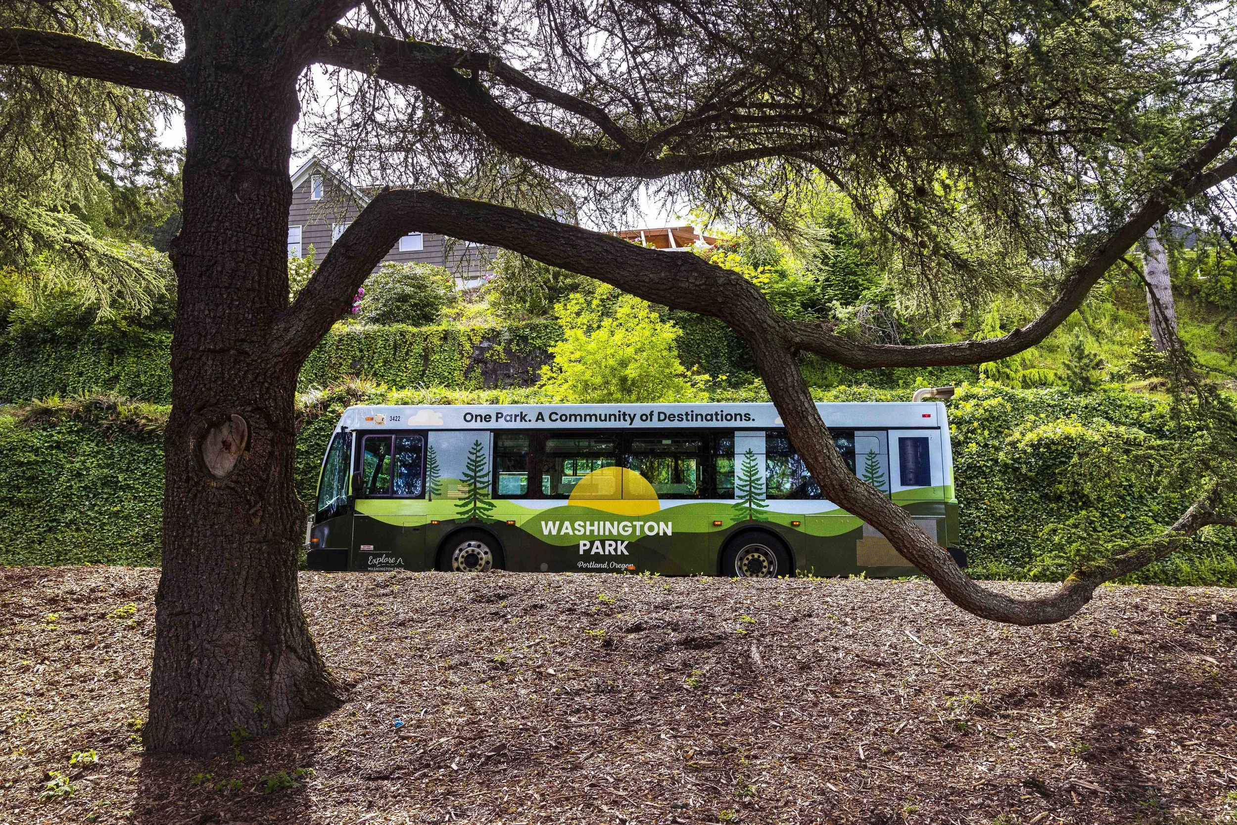 A Washington Park shuttle bus with a nature-themed design parked among trees, with a house and construction visible in the background.