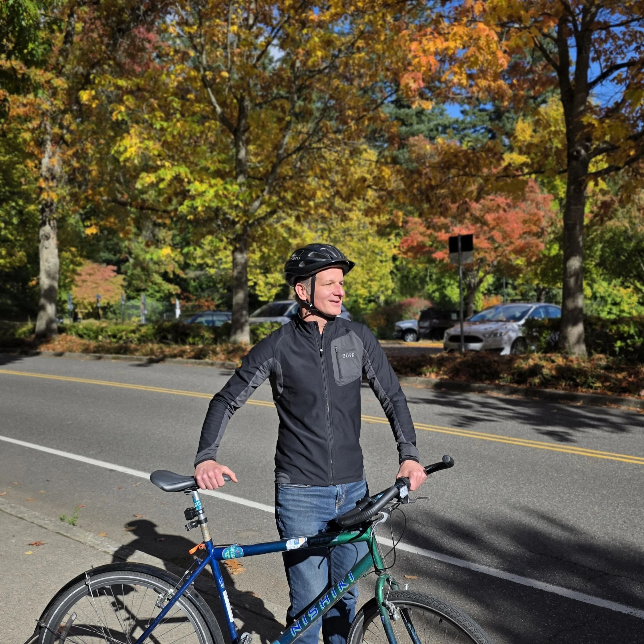 A cyclist wearing a helmet stands with his road bike on a tree-lined Washington Park road in autumn