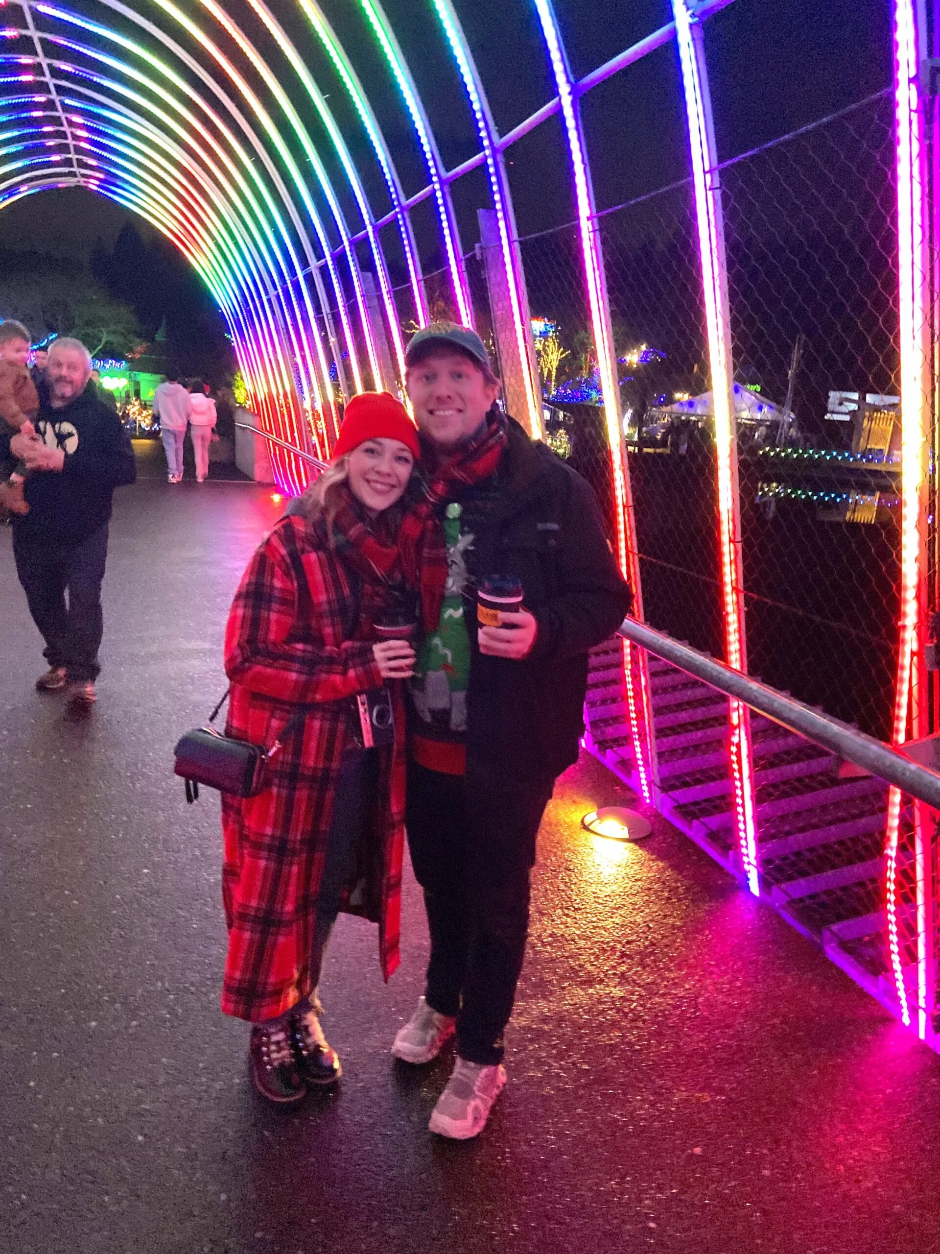 A couple posing under a colorful illuminated rainbow arch tunnel at ZooLights at the Oregon Zoo