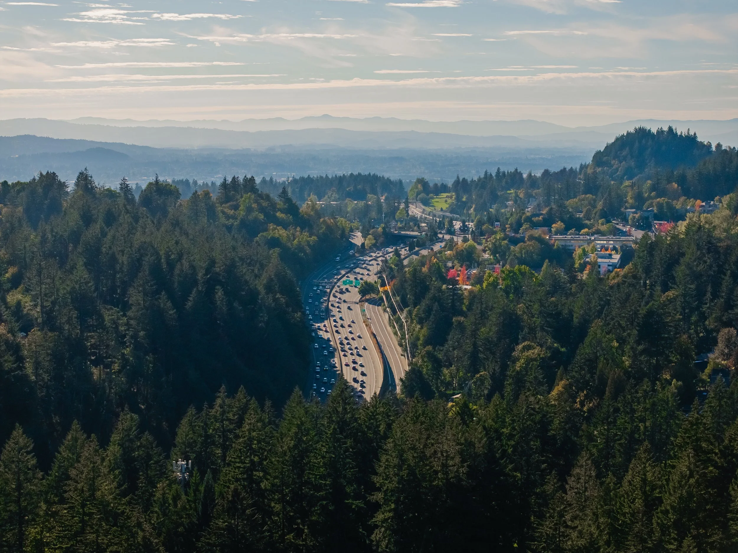 A winding highway through a forested mountainous area, with a town nearby and layers of distant mountain ranges under a partly cloudy sky.