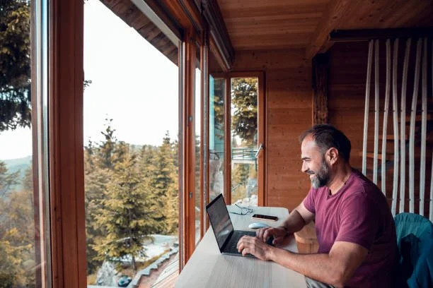 Man working on laptop in a wooden room with large windows showing trees and outdoors.