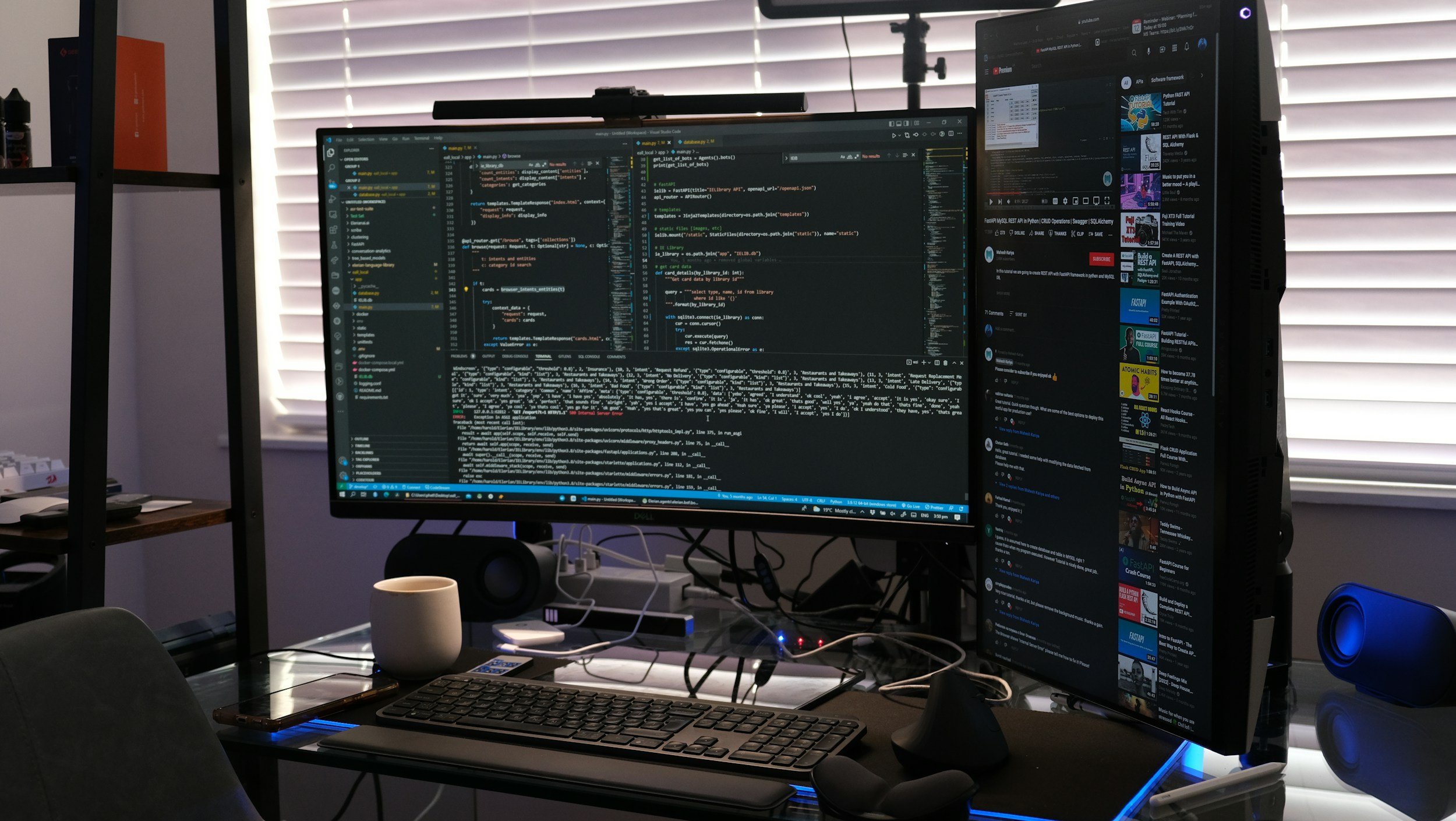 A dual-monitor computer setup on a desk in a room with closed blinds. The left screen displays a coding editor with lines of code, while the right screen shows a YouTube video playlist and comments. The desk has a keyboard, mouse, smartphone, a white mug, and various cables. A speaker is visible on the right side.