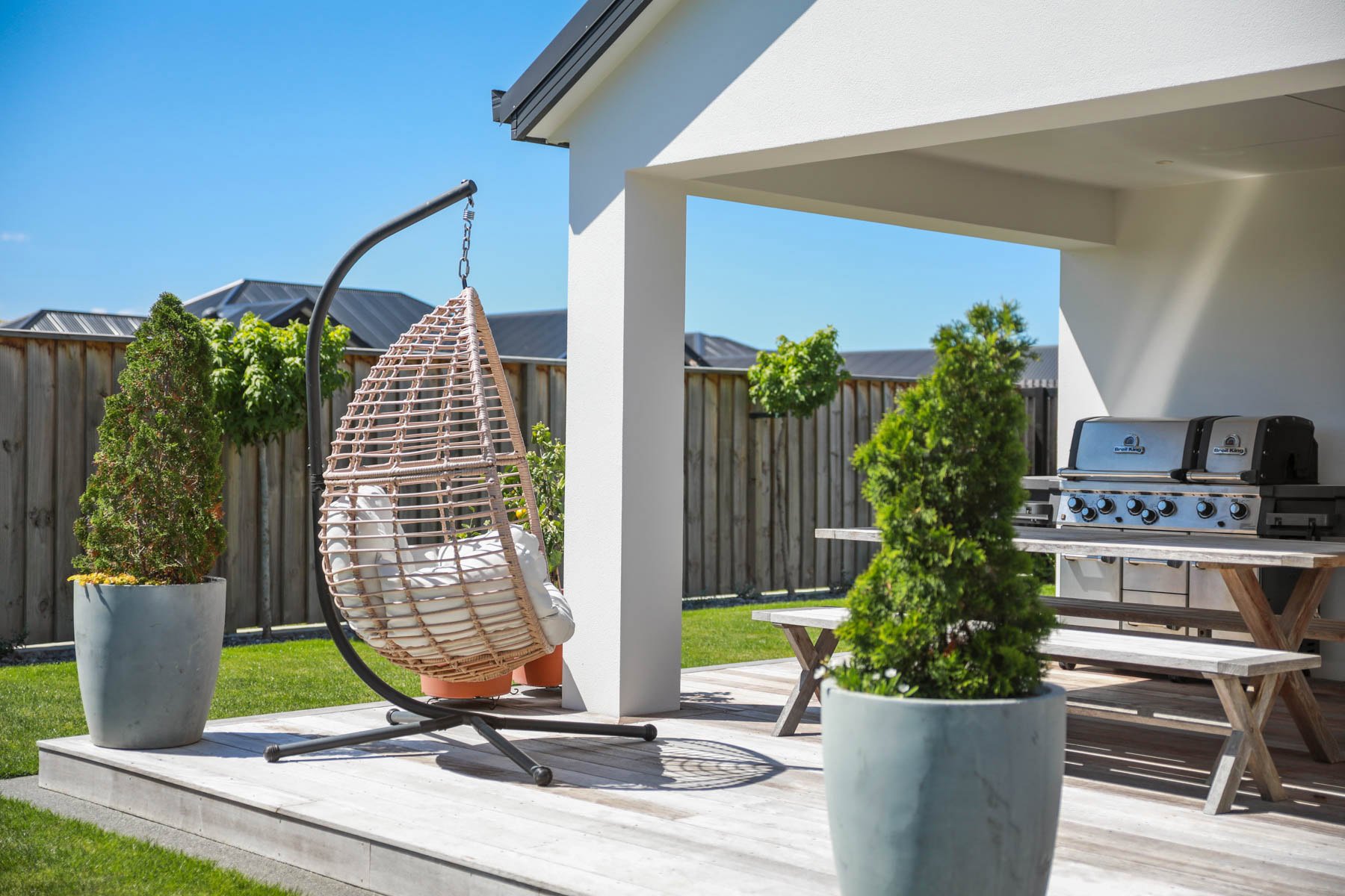 A backyard patio with a hanging wicker egg chair with white cushions, potted plants, a wooden picnic table, and a gas grill under a covered area on a sunny day.
