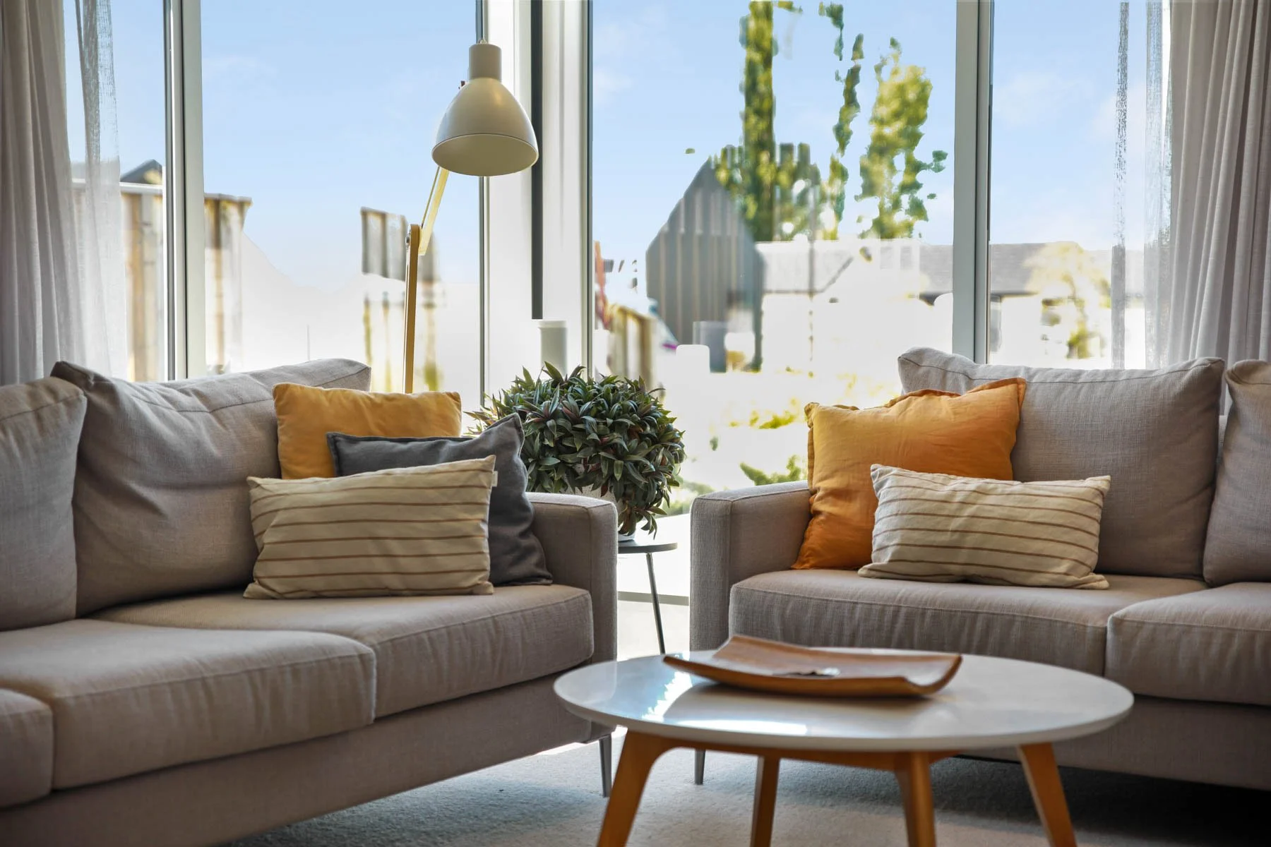 Living room with beige sofas, colorful pillows, a white round coffee table, a tall floor lamp, and large glass windows with curtains, overlooking a backyard.