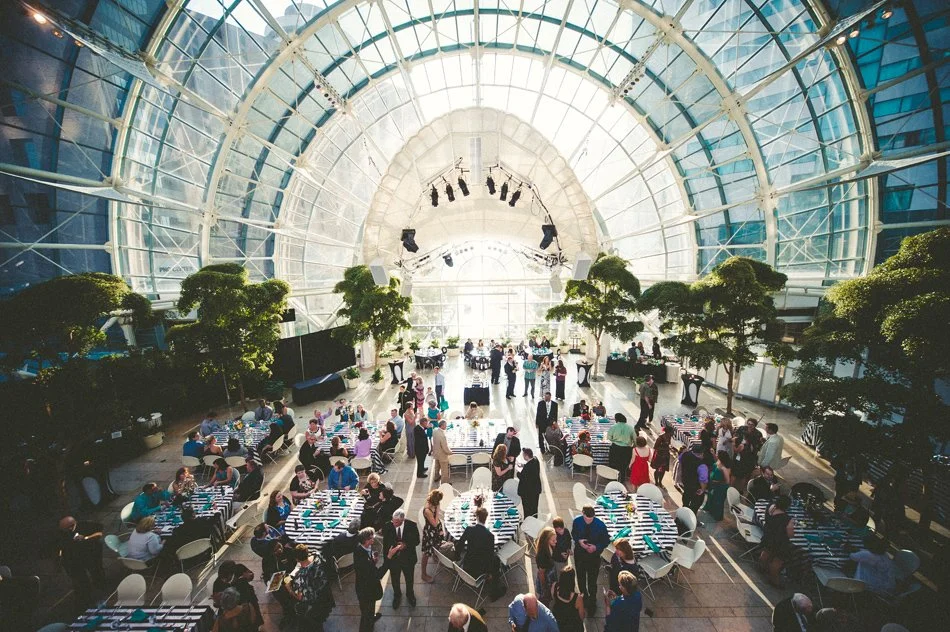 Indoor event space with a glass domed ceiling, decorated with green trees, hosting a large gathering of people dining at round tables with striped tablecloths.