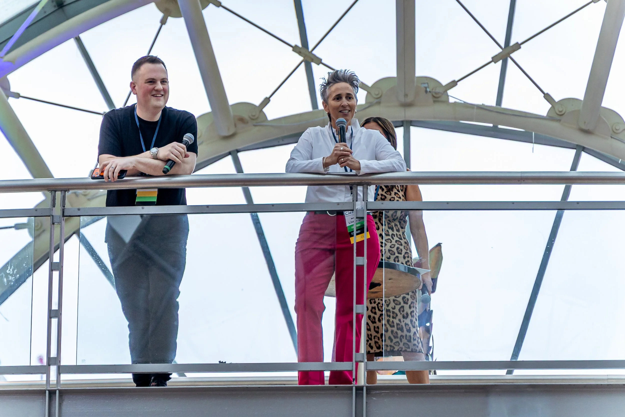 Three people standing on a balcony with glass railing, with large structural beams and sky in the background. The woman in the middle is speaking into a microphone, wearing a white shirt and red pants. The man on her left is holding a microphone and smiling, wearing a dark t-shirt. A third person with leopard print clothing is partially visible behind her.