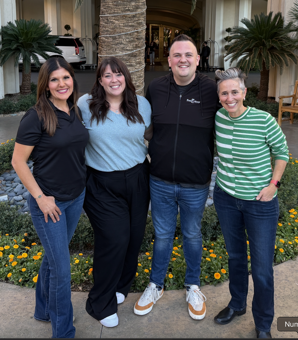 Four people standing outdoors in front of a tall palm tree and a building entrance, smiling at the camera.