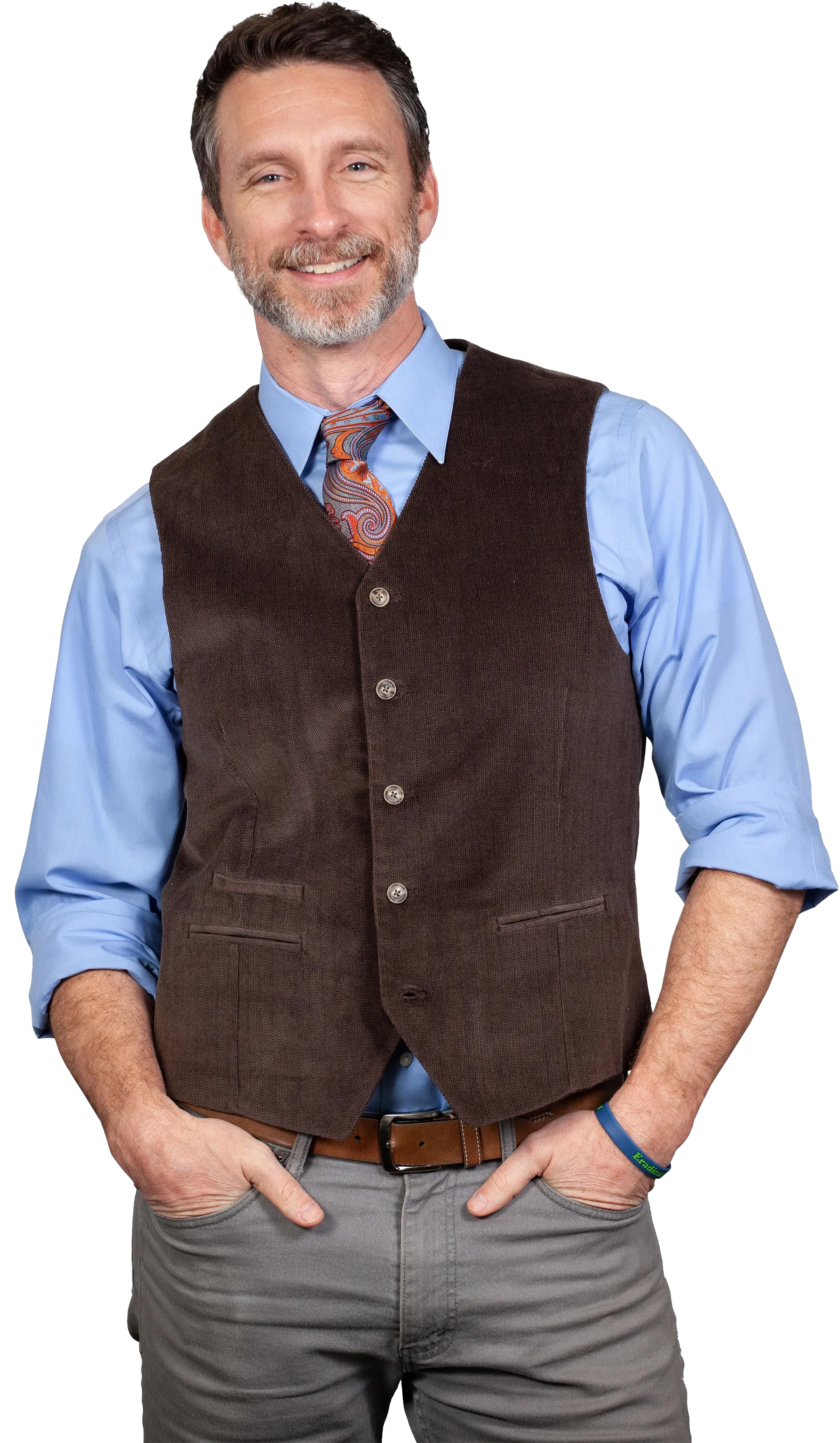 A middle-aged man with a beard and mustache, wearing a light blue dress shirt with the sleeves rolled up, a brown vest, a patterned tie, gray pants, and a brown belt, smiling and posing with hands in pockets.