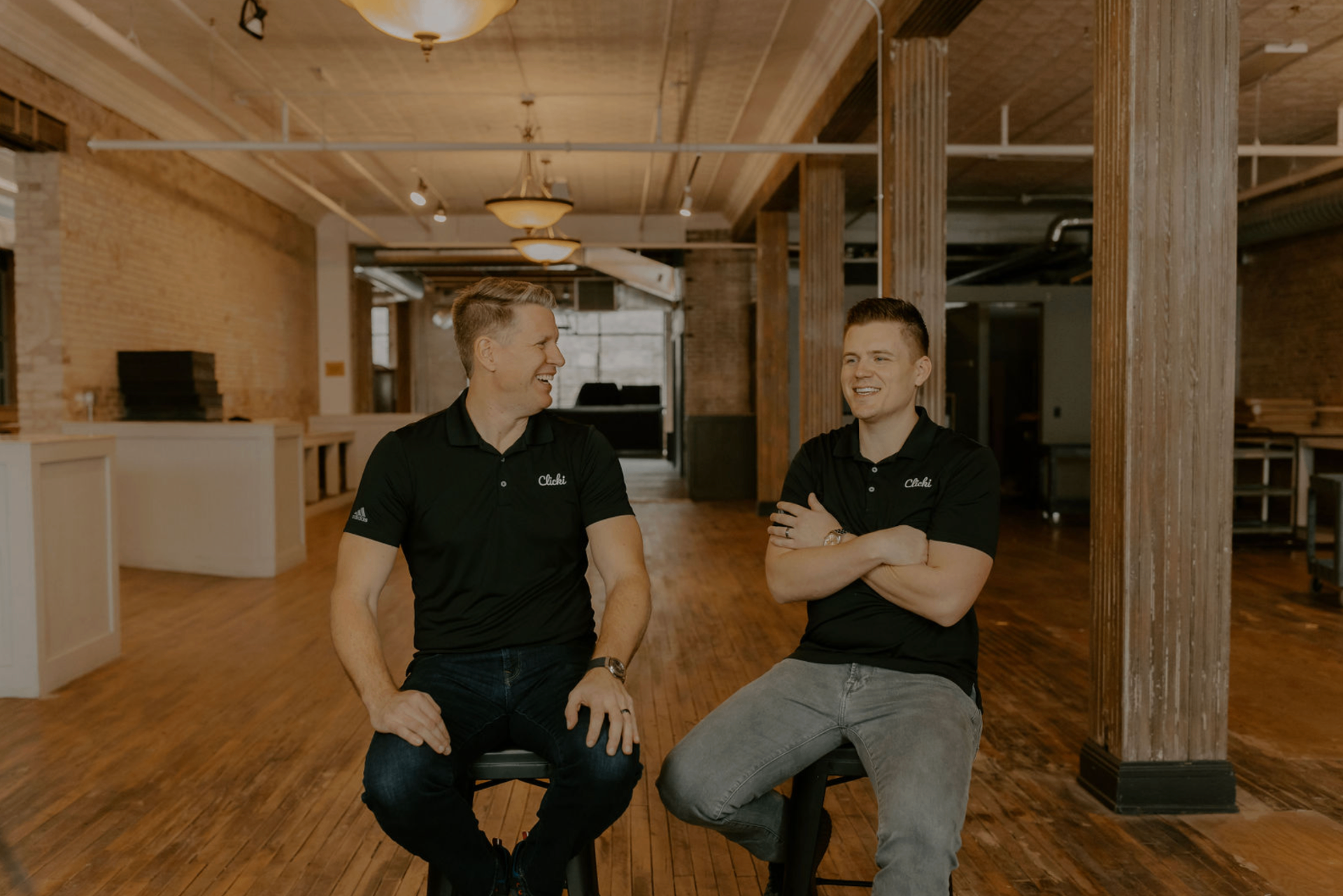 Two men sitting on chairs in a spacious, well-lit wooden interior, engaged in conversation, smiling, and laughing.