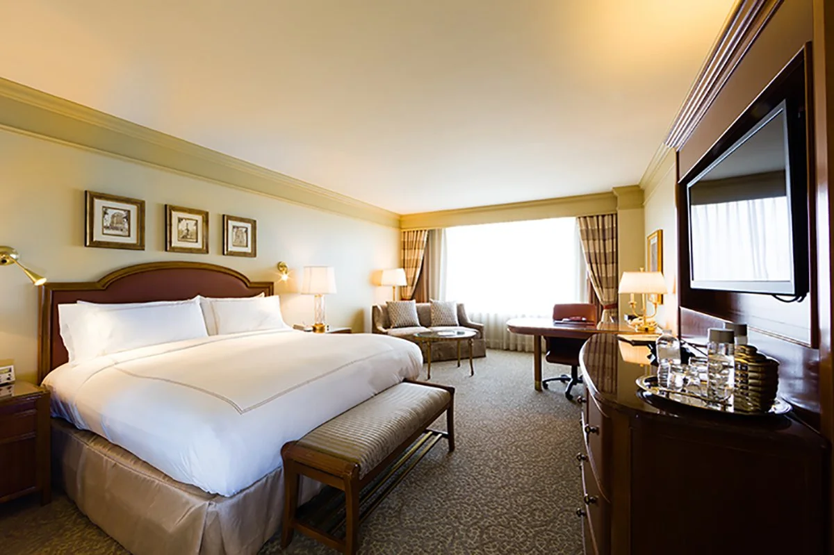 A spacious hotel room with a large bed, bedside tables with lamps, a sitting area near the window, a desk with a chair, and a large flat-screen TV mounted on the wall.