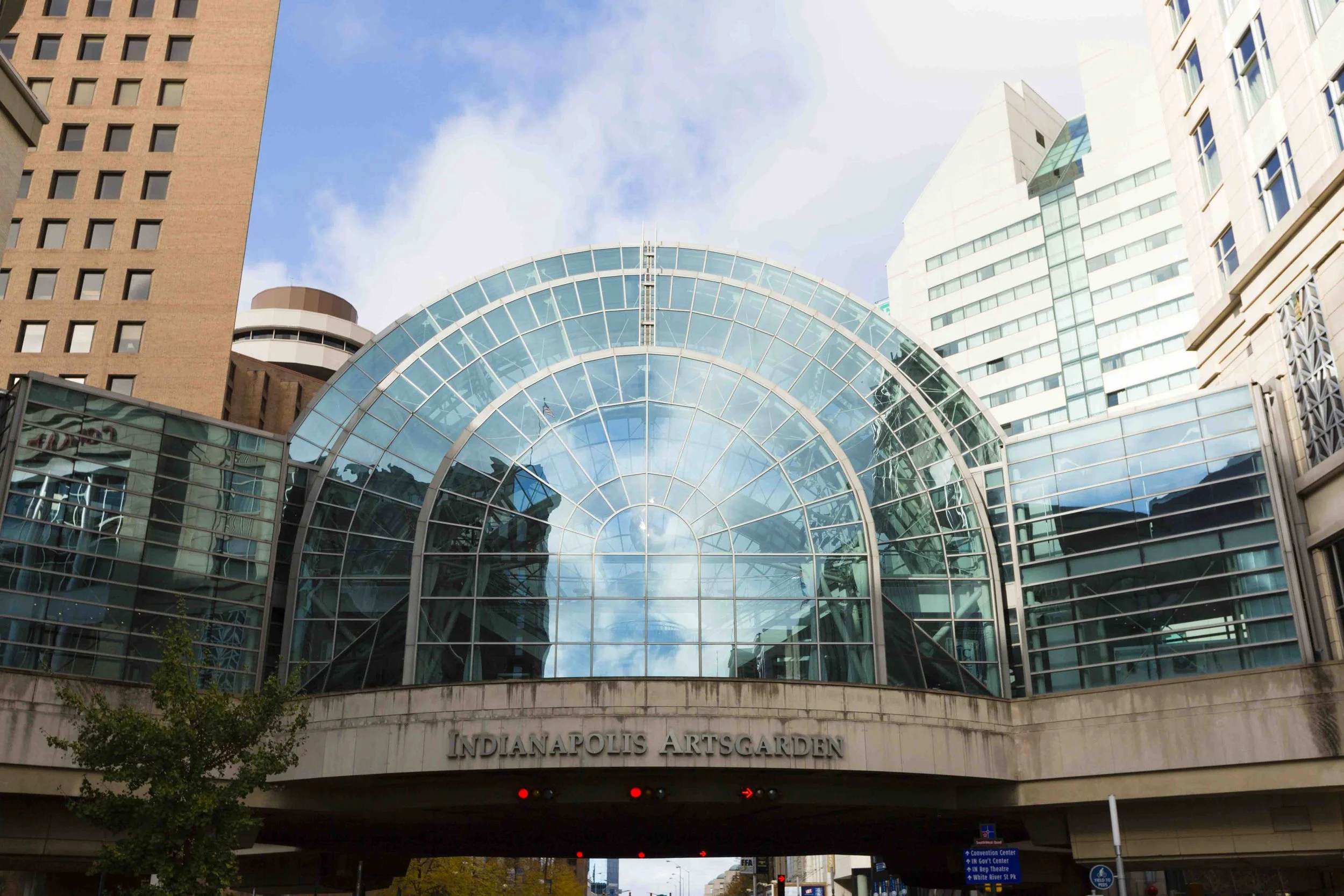 The Indianapolis Artsgarden, a glass-domed archway connecting buildings in Indianapolis, Indiana, with surrounding high-rise buildings under a partly cloudy sky.