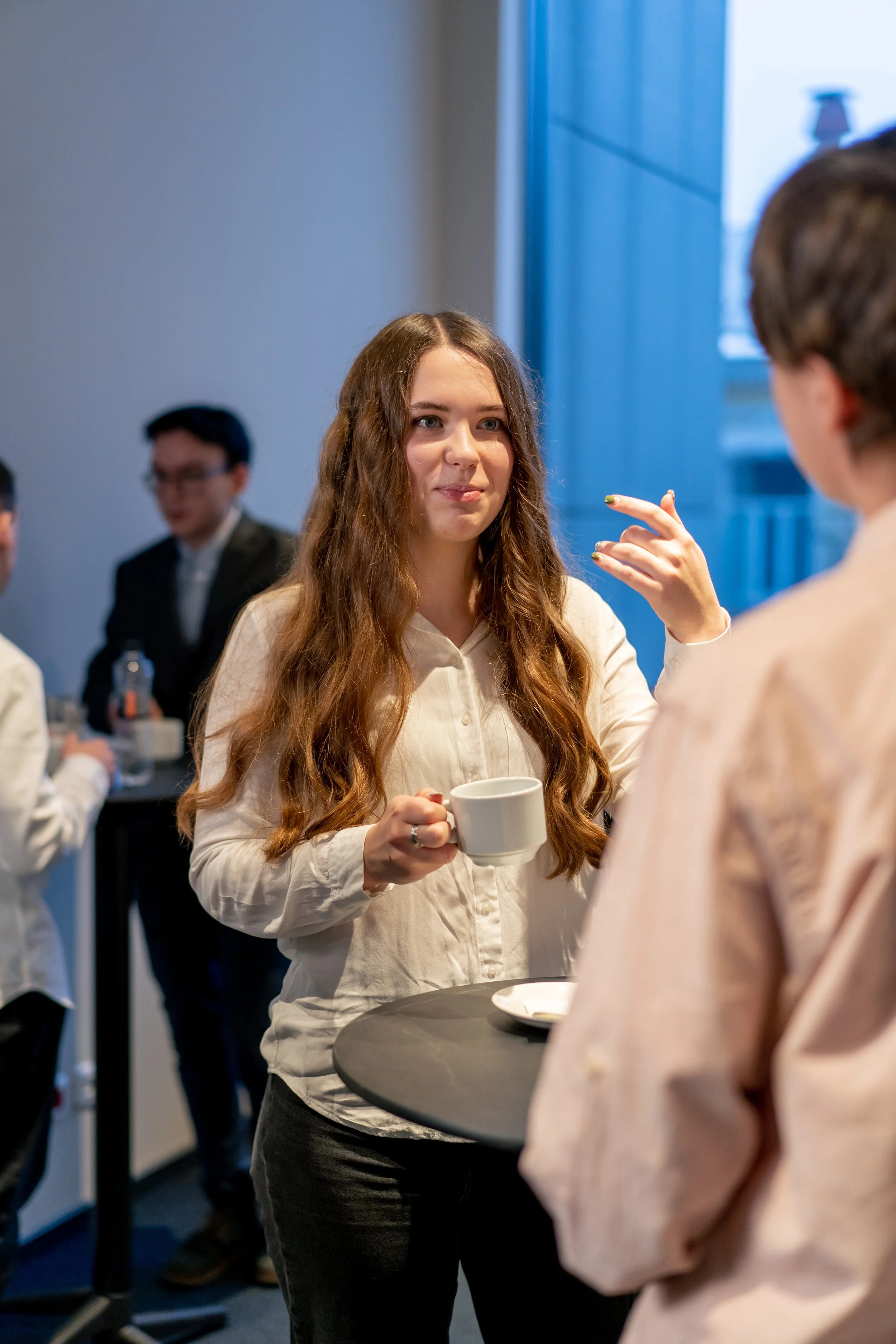 A young woman with long wavy hair holding a coffee mug, engaging in a conversation during a social gathering or meeting.