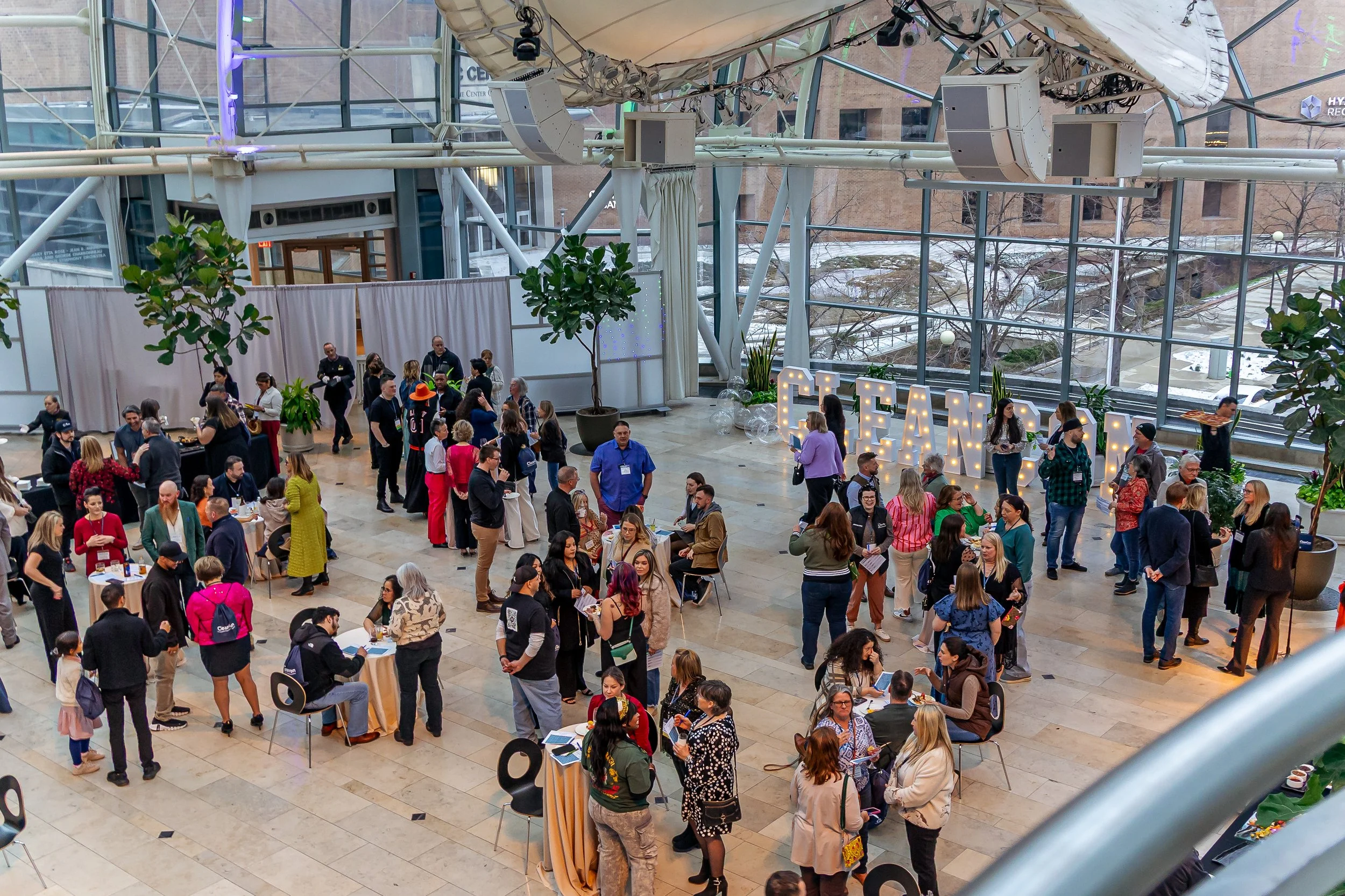 A busy indoor event with many people gathered around tables, some standing and some sitting, in front of large illuminated letters spelling 'FEAR'. The space has a high glass ceiling with metal framework, and potted plants are scattered around. There are people talking and networking, and a small stage or presentation area is visible in the background.