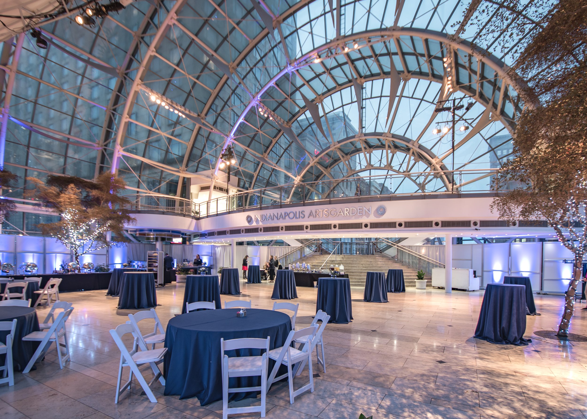 Elegant event space inside a glass-enclosed building with high arched ceiling, decorated for a formal gathering with tables covered in dark blue tablecloths, white chairs, and string lights on trees.