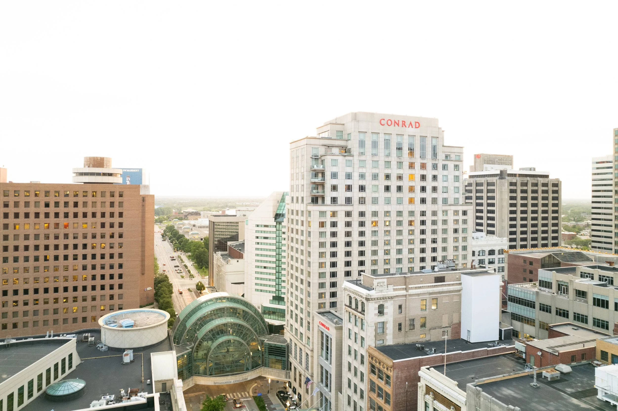 A cityscape view of tall buildings with one labeled 'CONRAD' and a modern glass structure at the bottom, with a street and greenery in the background.