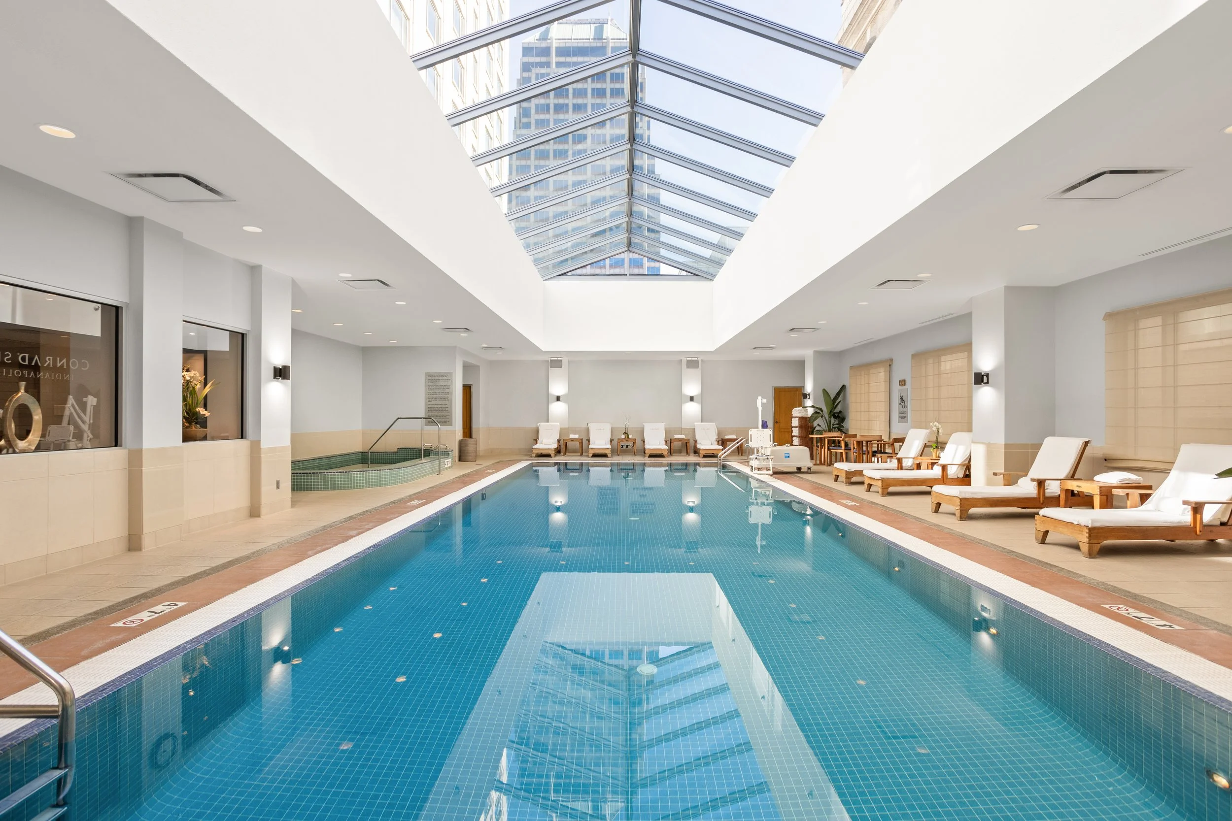 Indoor swimming pool area with lounge chairs along the sides under a glass ceiling with a cityscape view.