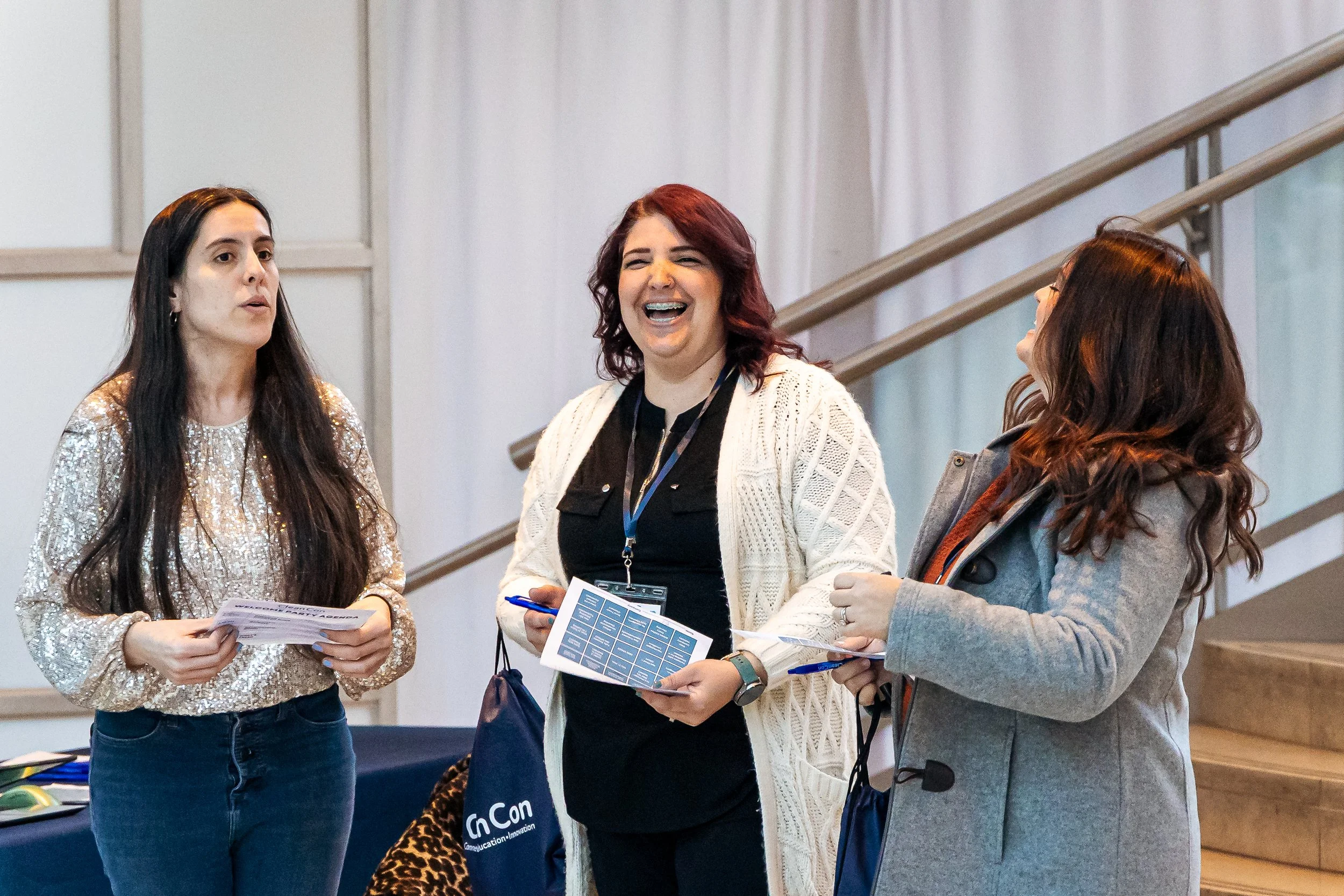 Three women standing and talking at an indoor event, holding informational pamphlets. One woman is wearing a shiny, gold-colored top, the second has red hair and a white cardigan, and the third is wearing glasses and a gray jacket.