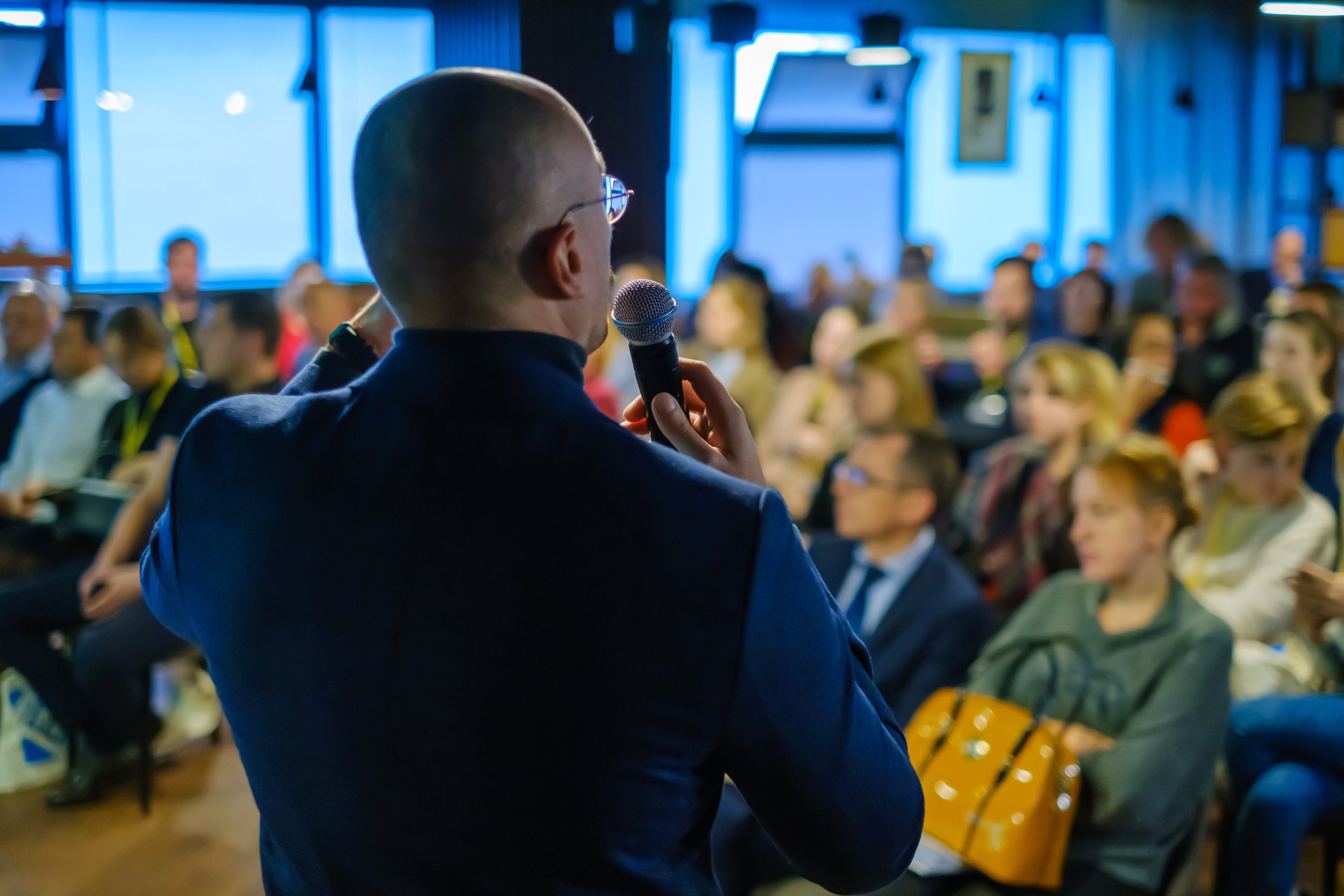A man holding a microphone and speaking to an audience during a conference or seminar, with the audience members seated and listening attentively.
