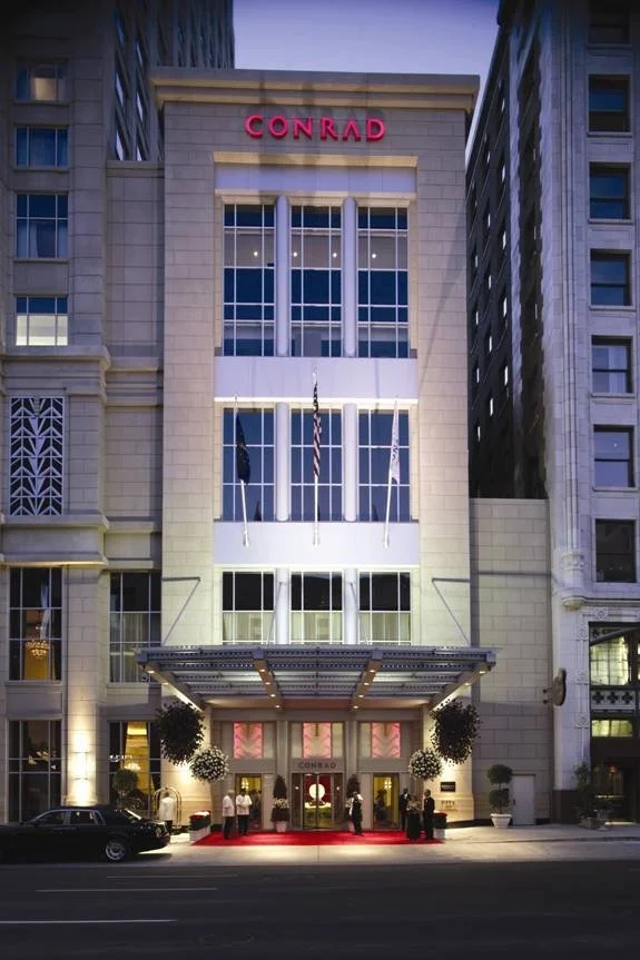 Night view of the Conrad hotel building with a red illuminated sign at the top, glass entrance doors, potted plants, and people gathered outside.