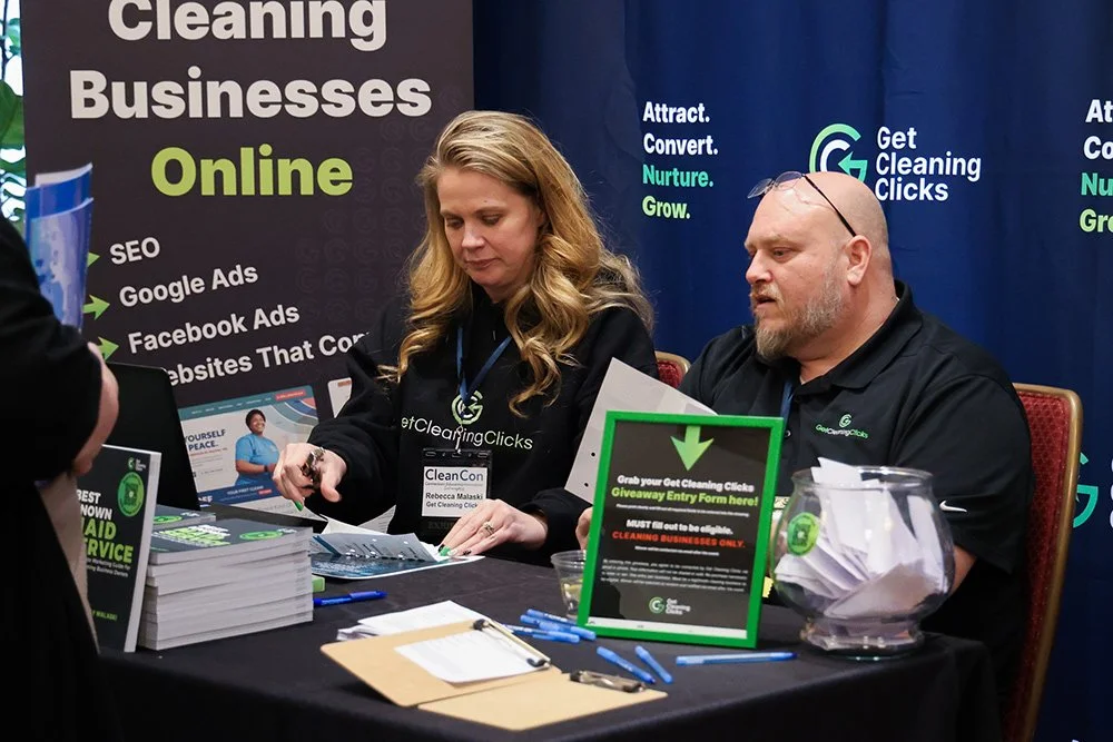 Two event staff members sitting at a table at a conference, with promotional materials about online cleaning businesses and a sign promoting a cleaning click giveaway entry.