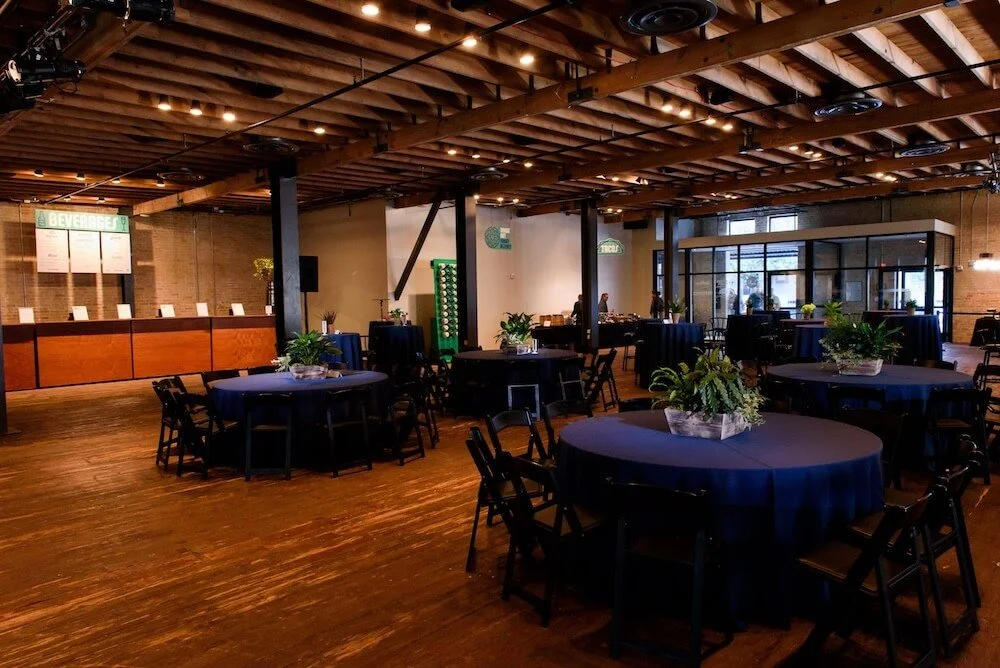 Indoor event space with round tables covered in navy blue tablecloths and potted plants, a stage area with a check-in counter, and a glass-enclosed room in the background.