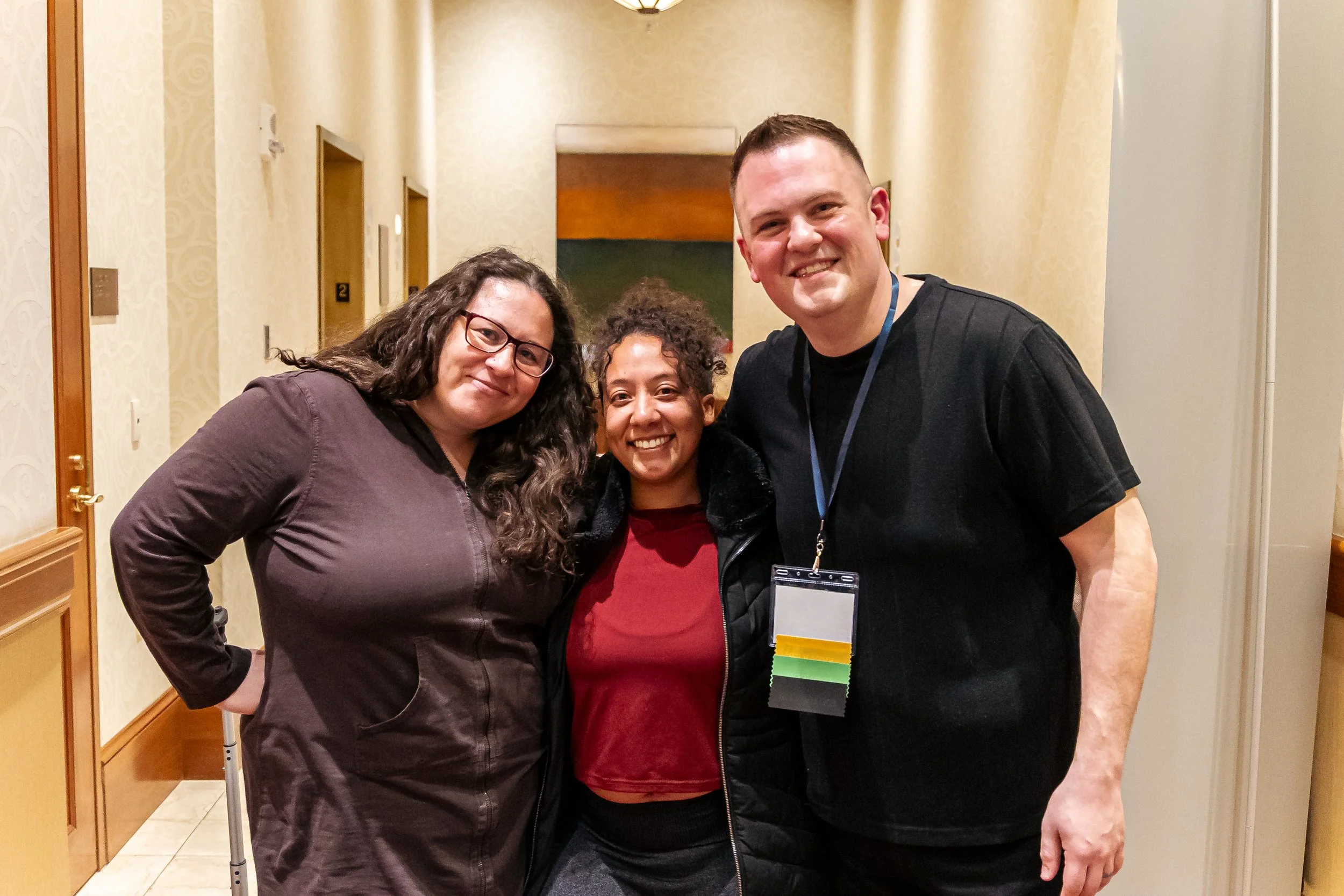 Three women and one man smiling and posing together indoors, with the man wearing a conference badge and a multicolored ribbon.