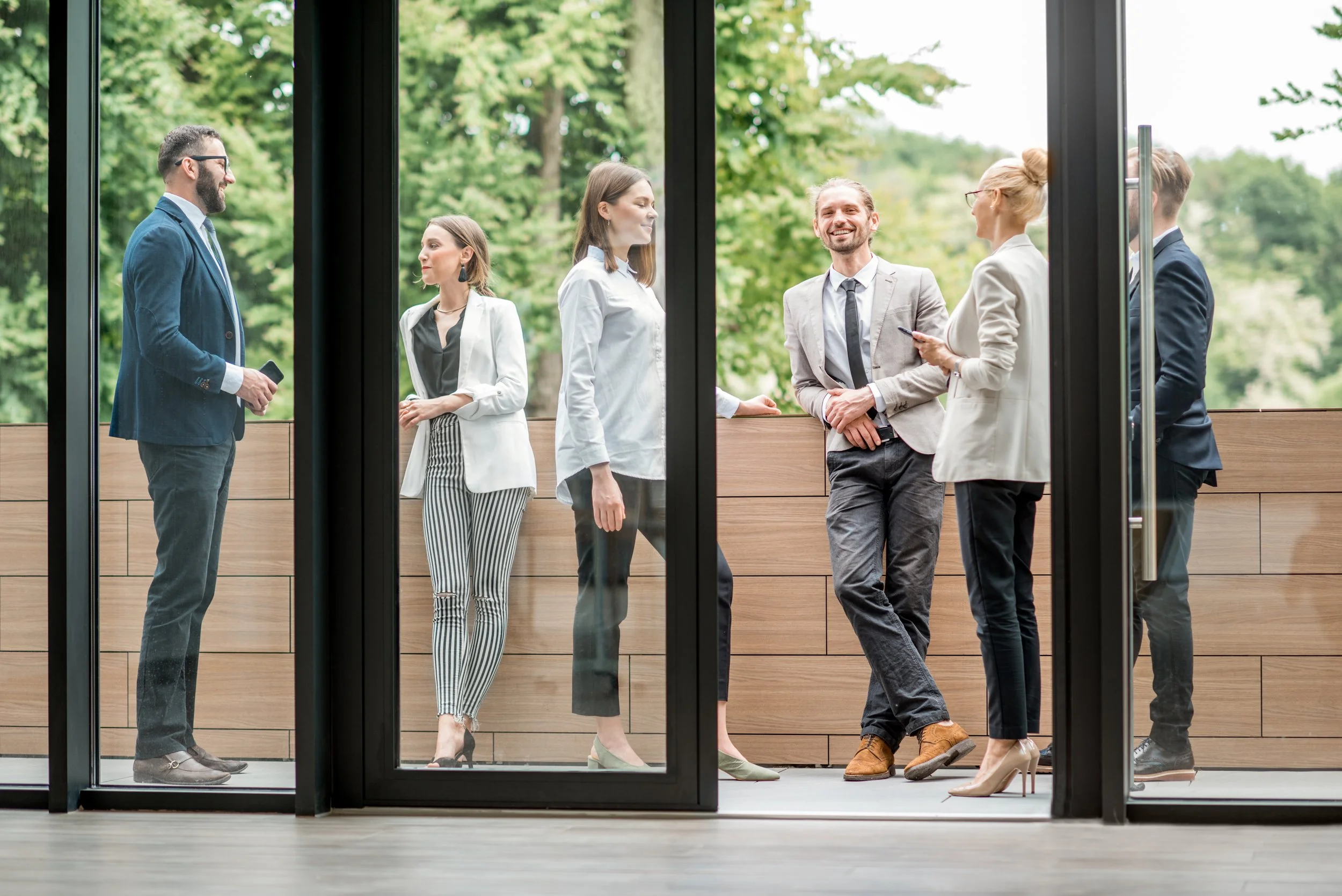 Group of six professionals in business attire standing on an outdoor balcony engaged in conversation, with trees in the background.