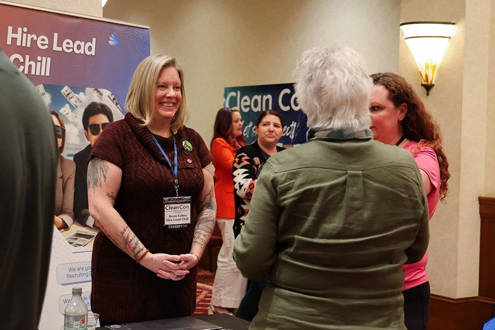 A woman with blonde hair and tattoos, wearing a dark red dress and a name badge, is smiling and talking to an older person with white hair and a green jacket at a conference or event. Several people are in the background, and banners with text like 'Hire Lead Chill' and 'Clean Co' are visible.